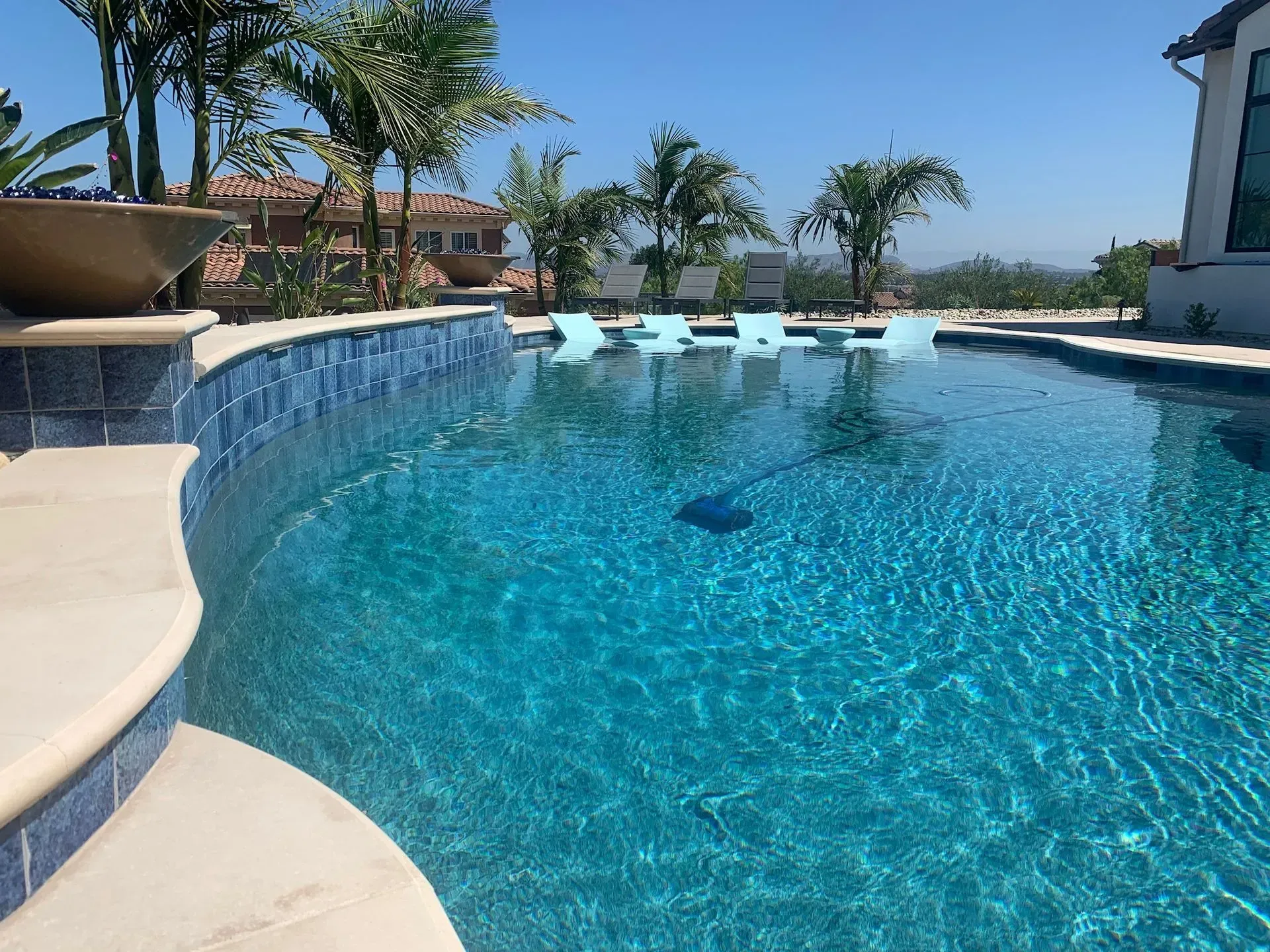 Blue tiled swimming pool with palm trees, and partial view of a house on a sunny day.