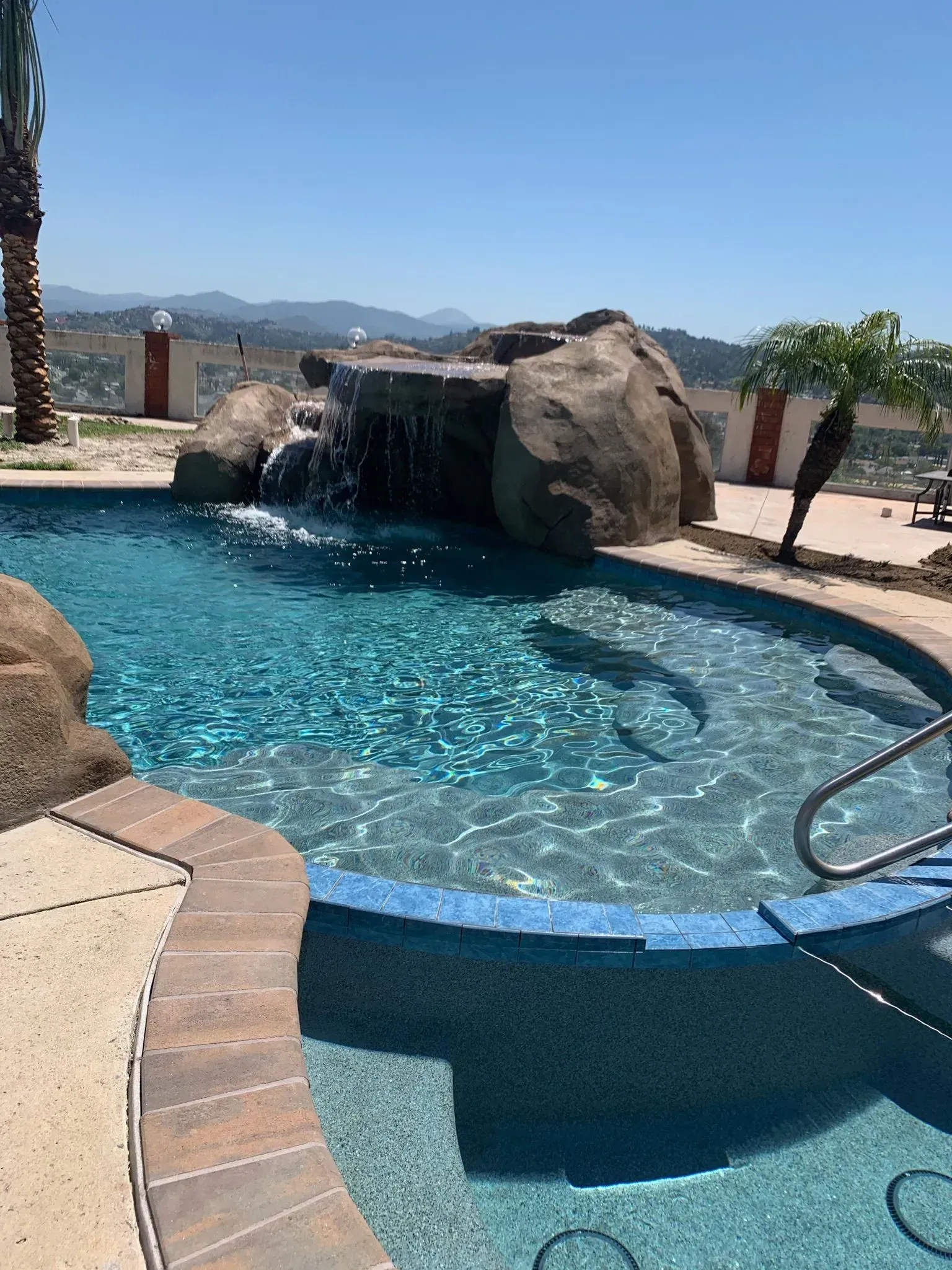 Pool with a waterfall feature and a view of distant hills on a sunny day.