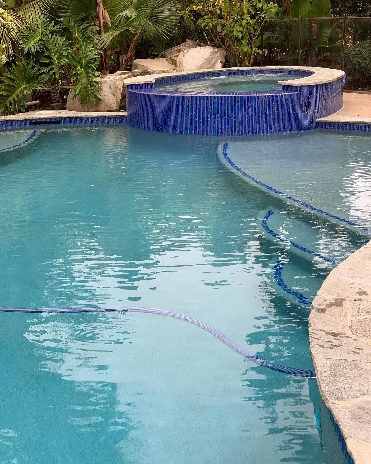 Blue-tiled pool and hot tub with waterfall feature, surrounded by stone and lush greenery.