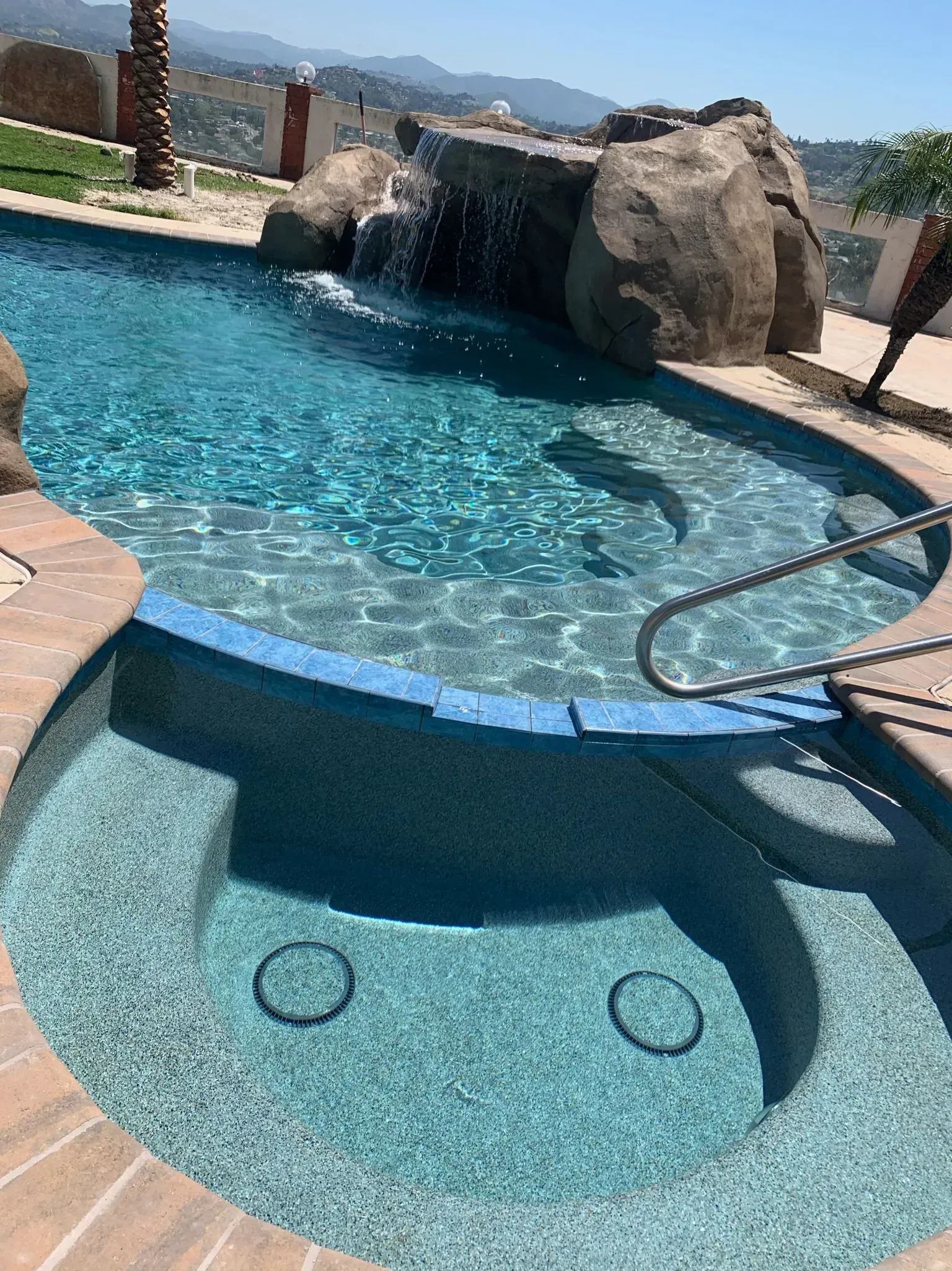 Pool with a waterfall, spa, and tan-colored surrounding, overlooking a mountainous landscape.