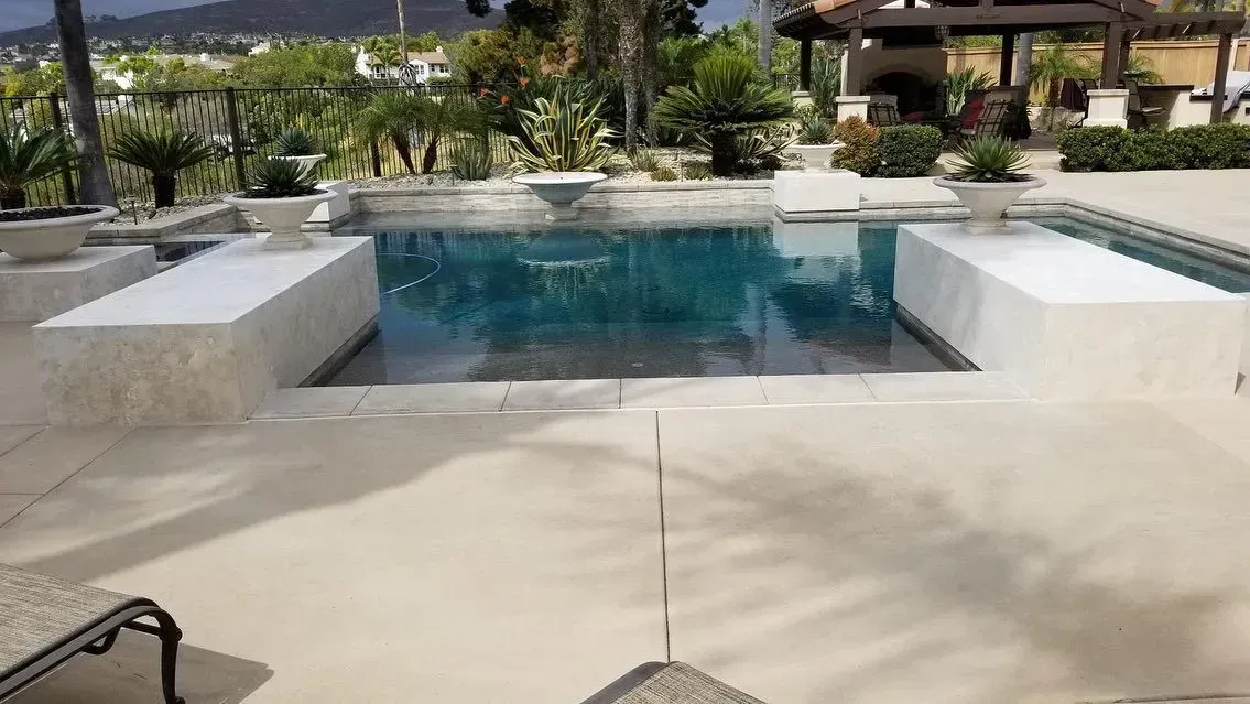 Swimming pool with white built-in benches, surrounded by patio. Green landscaping and sunny setting.
