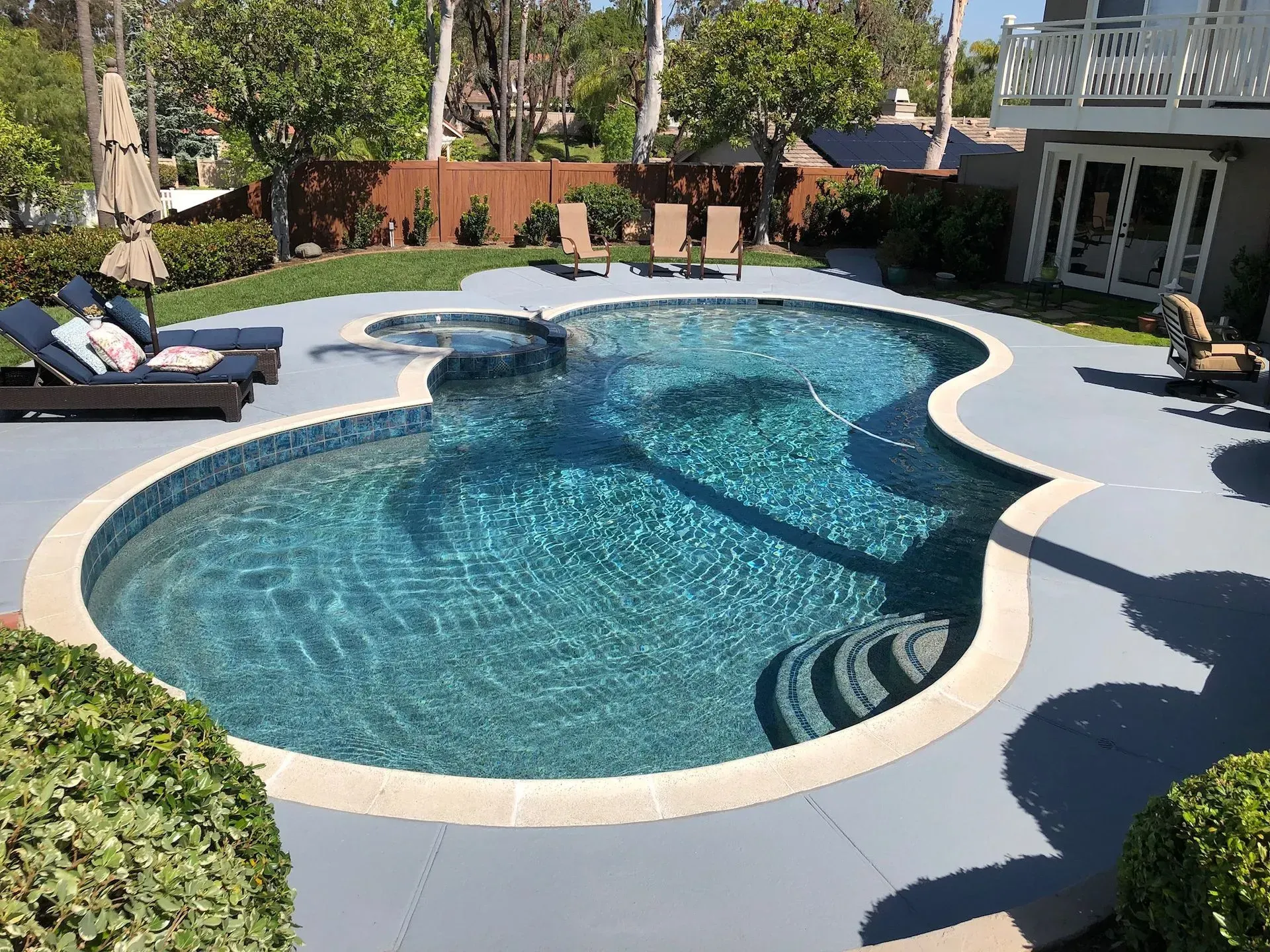 A backyard pool with a hot tub and lounge chairs. Blue water and gray concrete surround the pool.