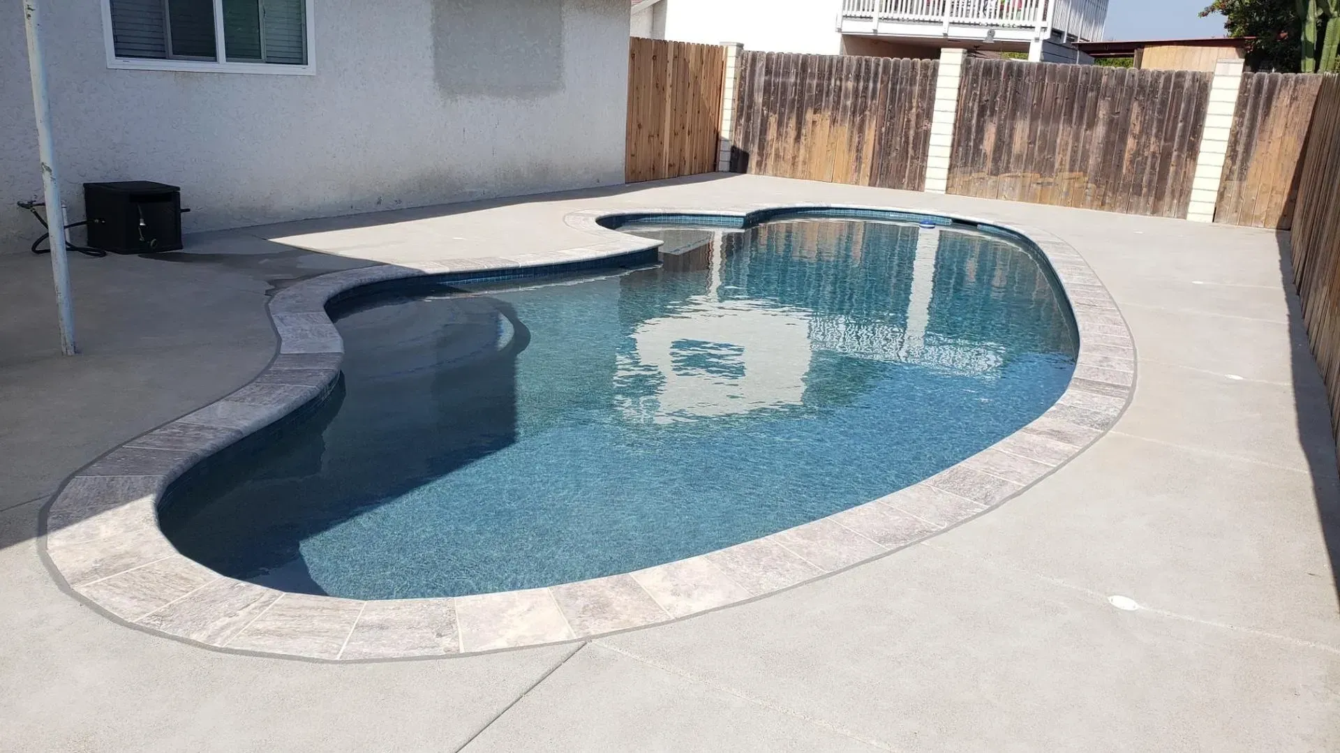 Pool with blue water and stone coping in a backyard with concrete patio.