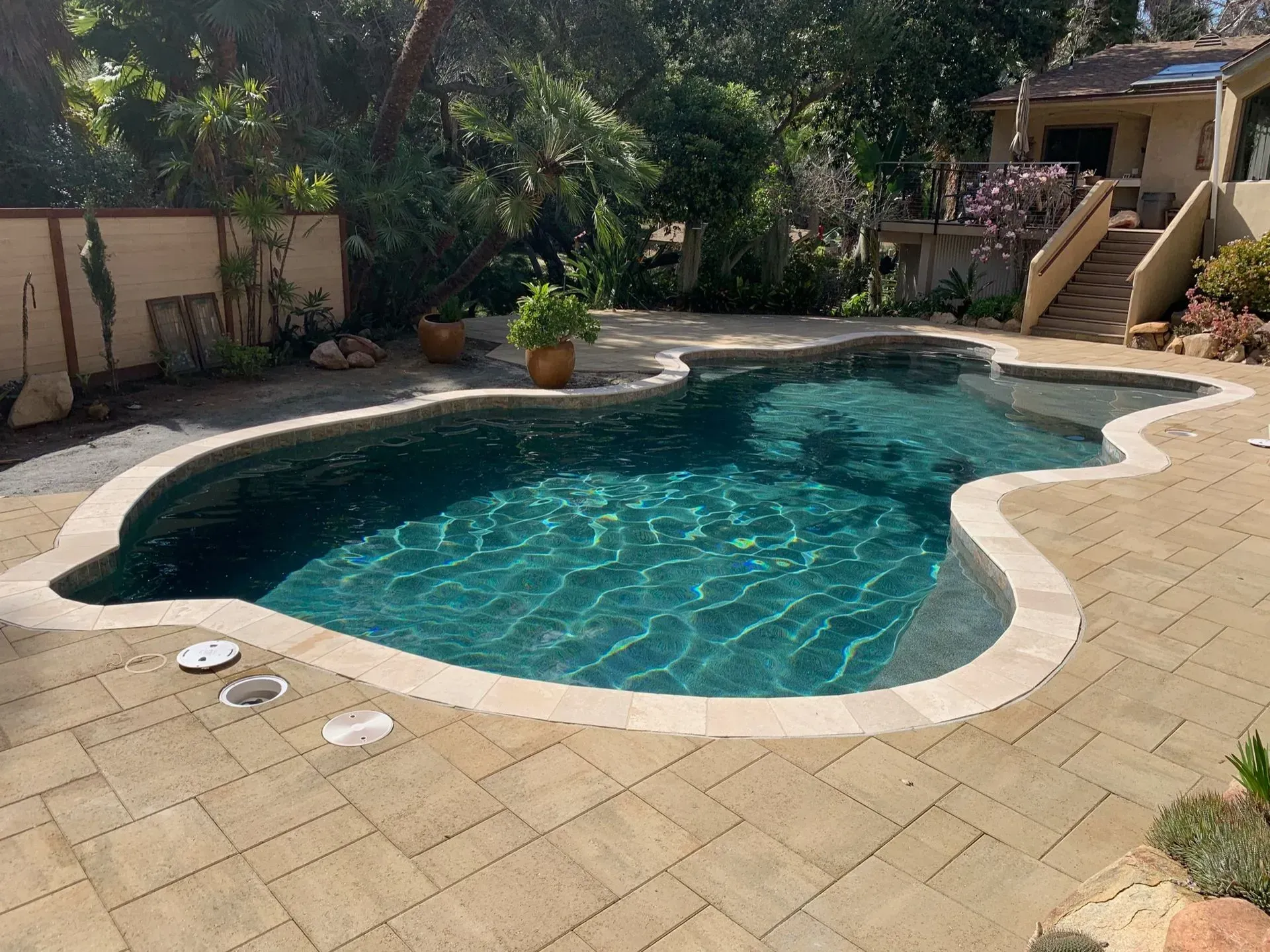 Swimming pool with turquoise water, surrounded by stone patio, trees, and a house.