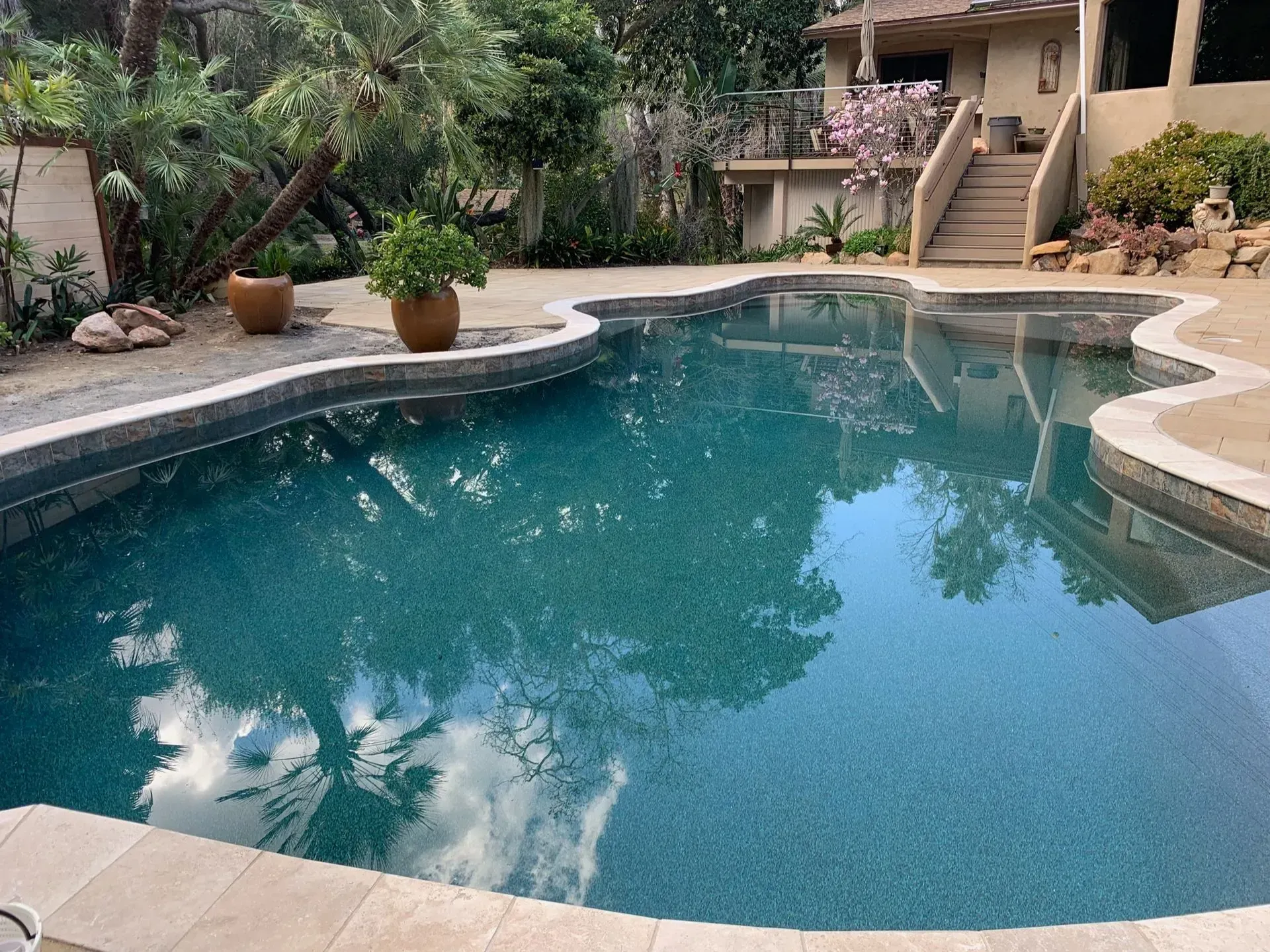 Pool with dark blue water surrounded by a light-colored patio. Trees and a house in the background.