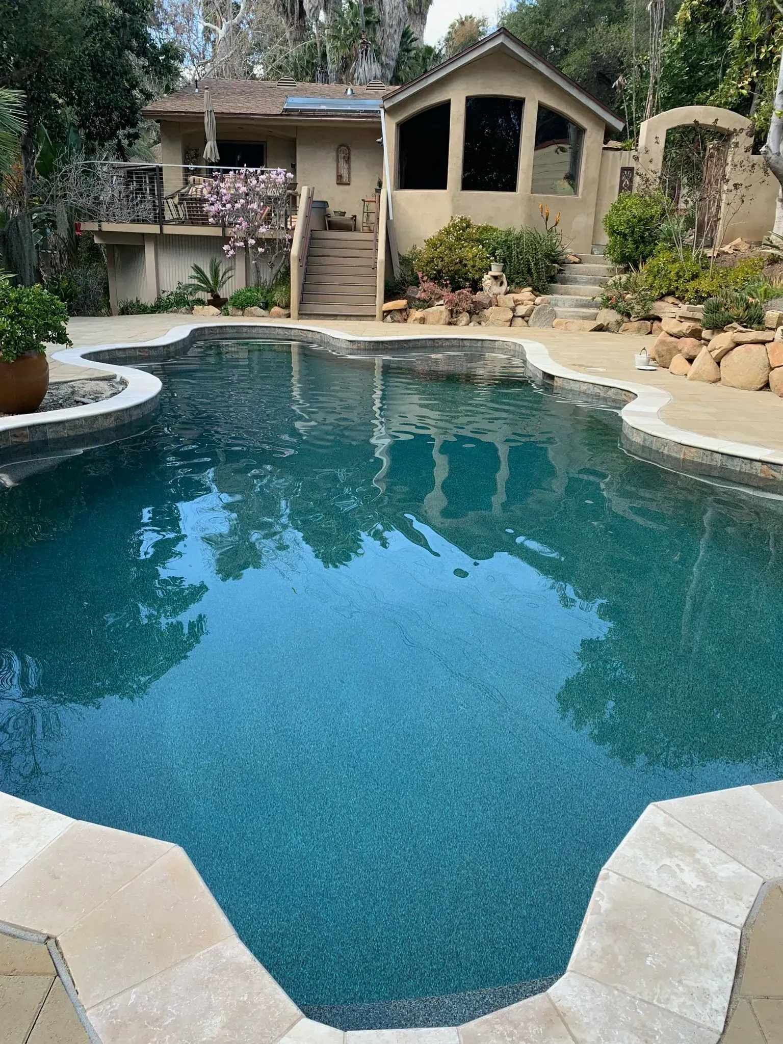 Swimming pool in front of a tan-colored house with a dark-roofed structure and lush landscaping.