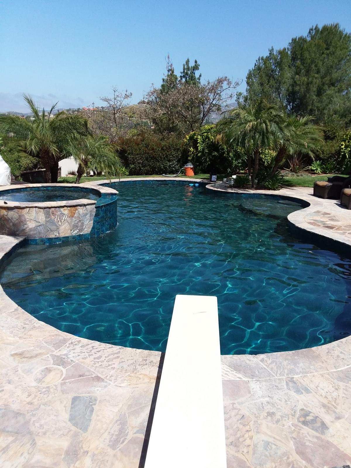 Swimming pool with diving board, surrounded by a stone patio, trees, and a spa. Blue water.