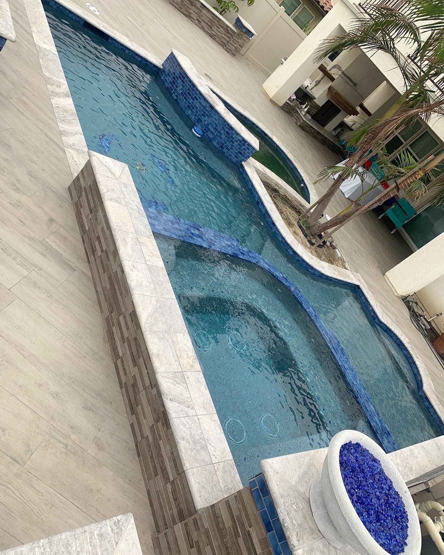 Swimming pool with blue tiles, stone border, and a small tree.