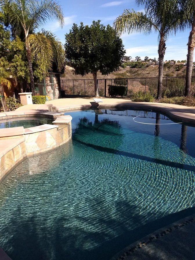 Pool and spa in a backyard on a sunny day. Palm trees and a tree reflect in the water.