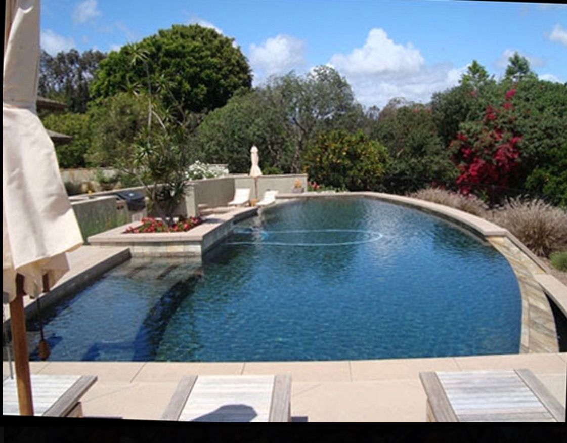 Swimming pool surrounded by stone patio and landscaping under a blue sky.