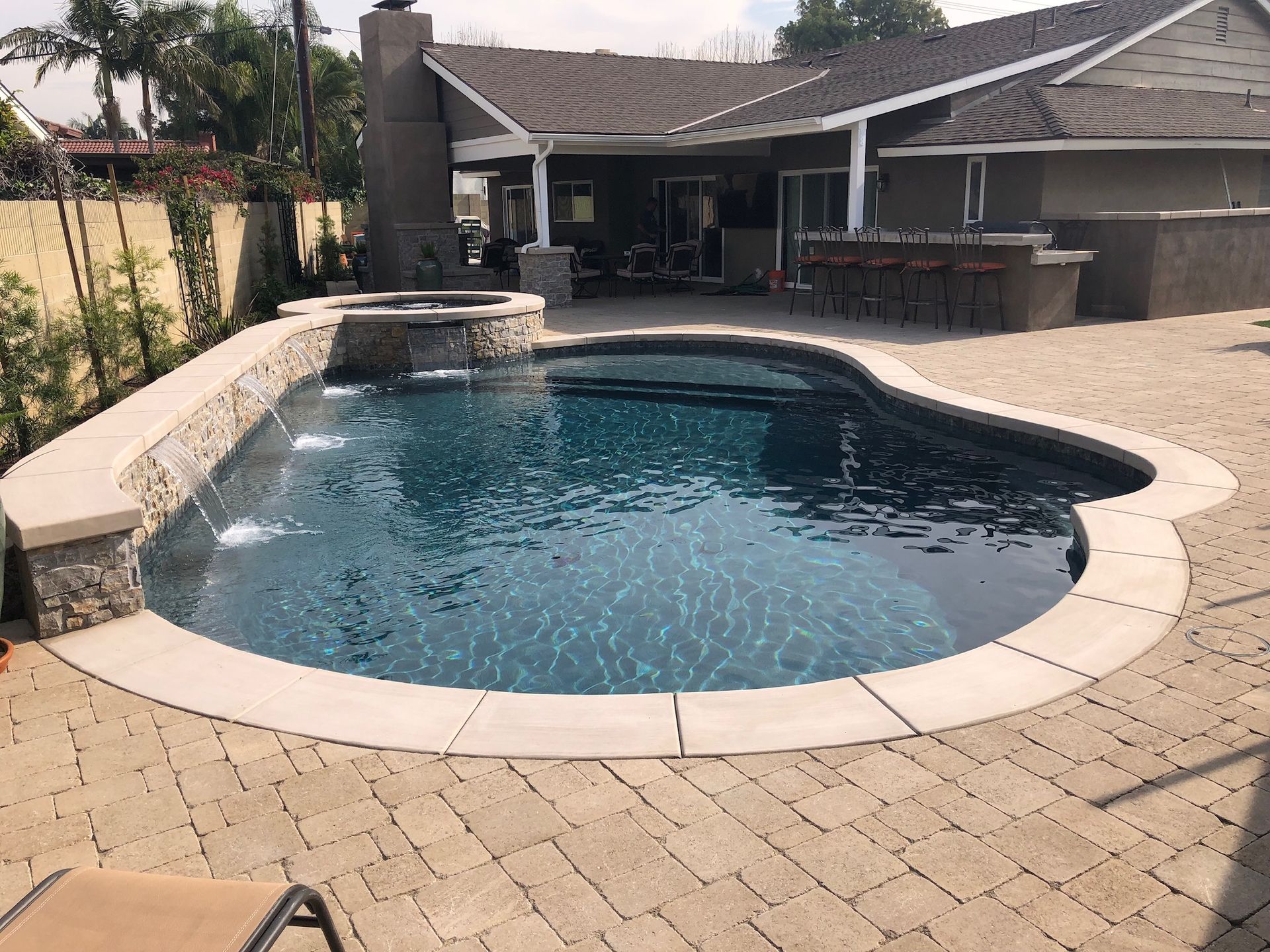 A swimming pool with stone coping and a hot tub, surrounded by a brick patio. The backyard setting includes a house.