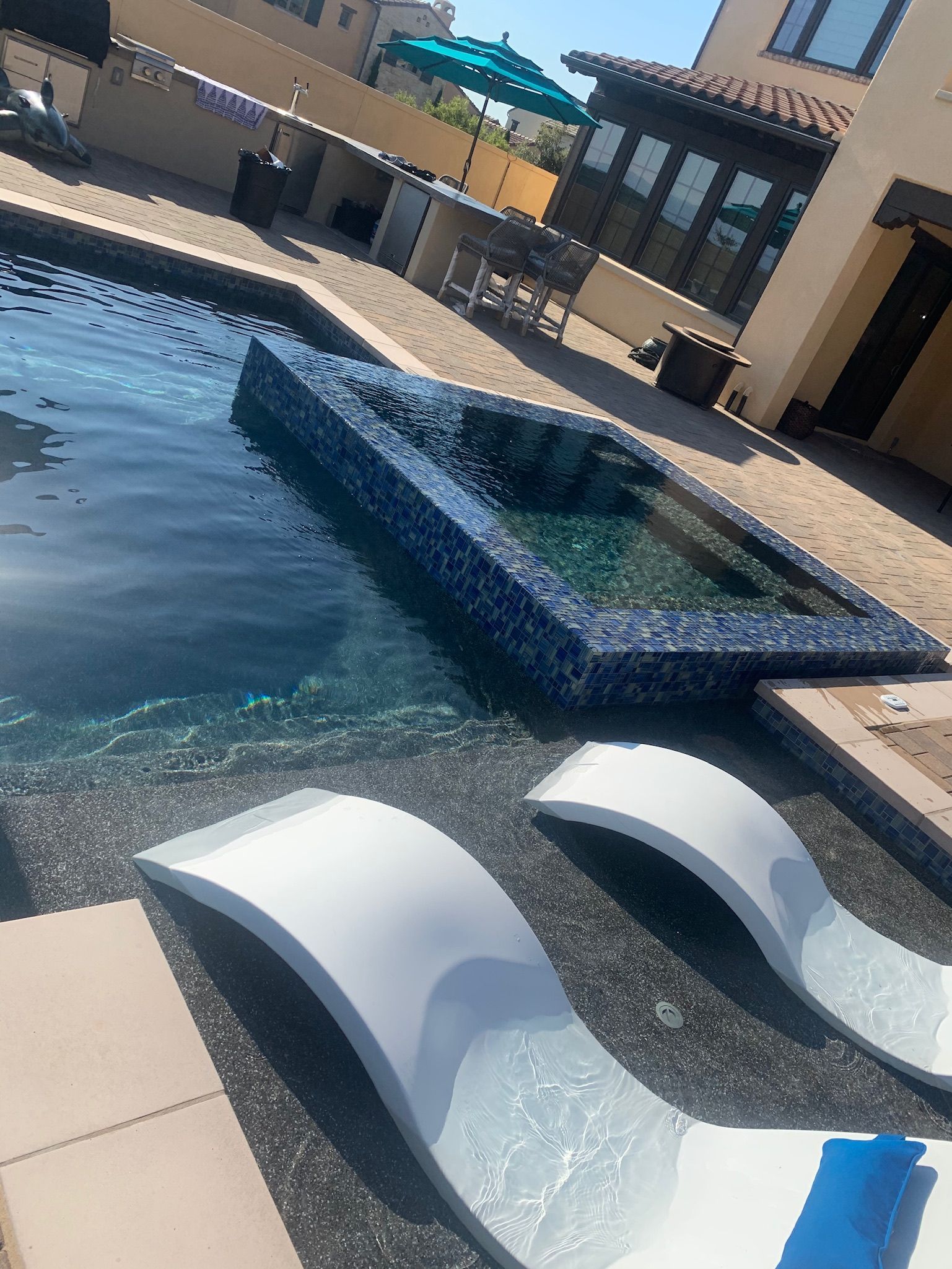 Pool with blue-tiled spa, white lounge chairs, and a patio, surrounded by a building with a blue umbrella.