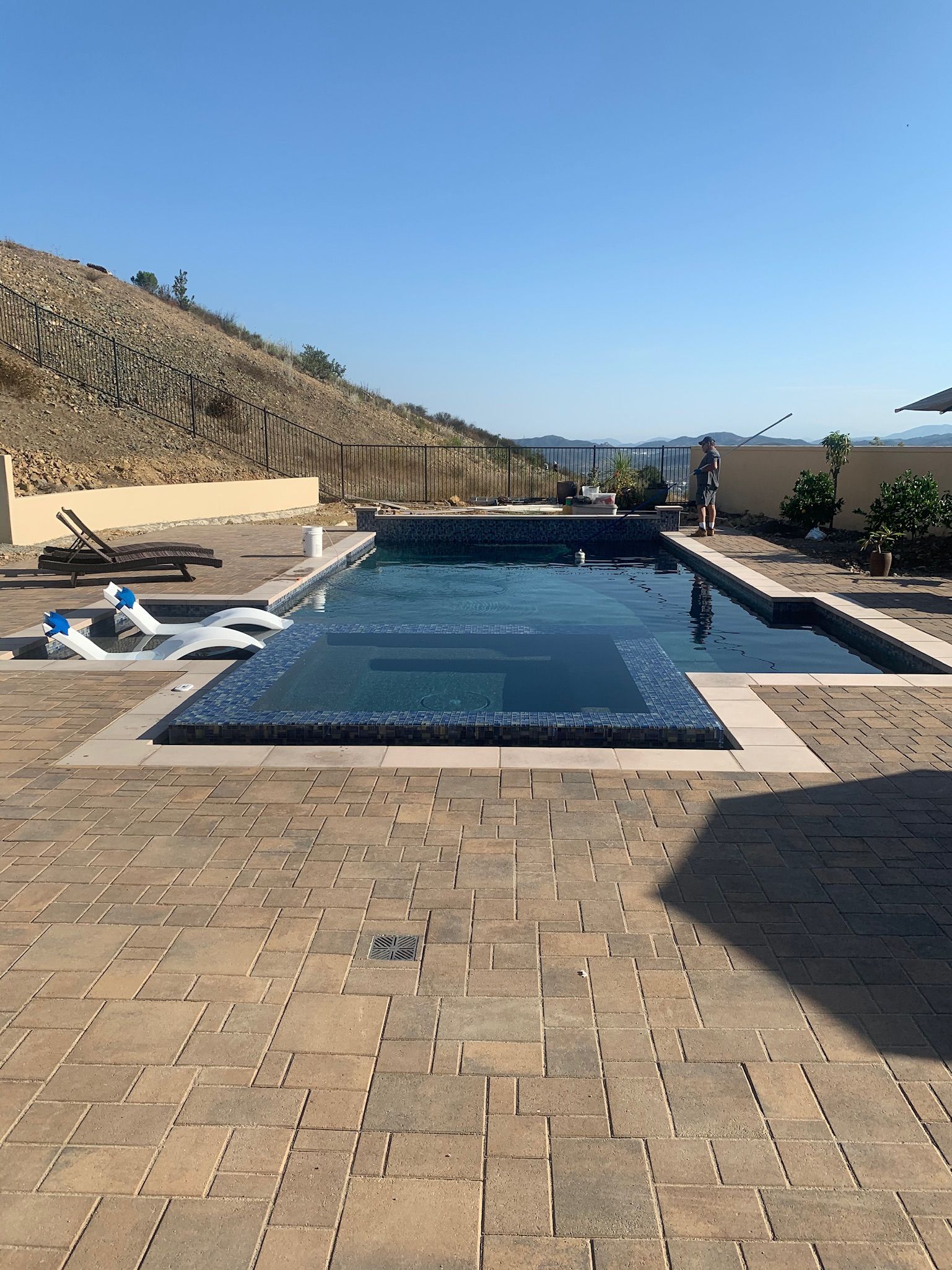 Backyard pool with stone patio, overlooking a hillside on a sunny day.