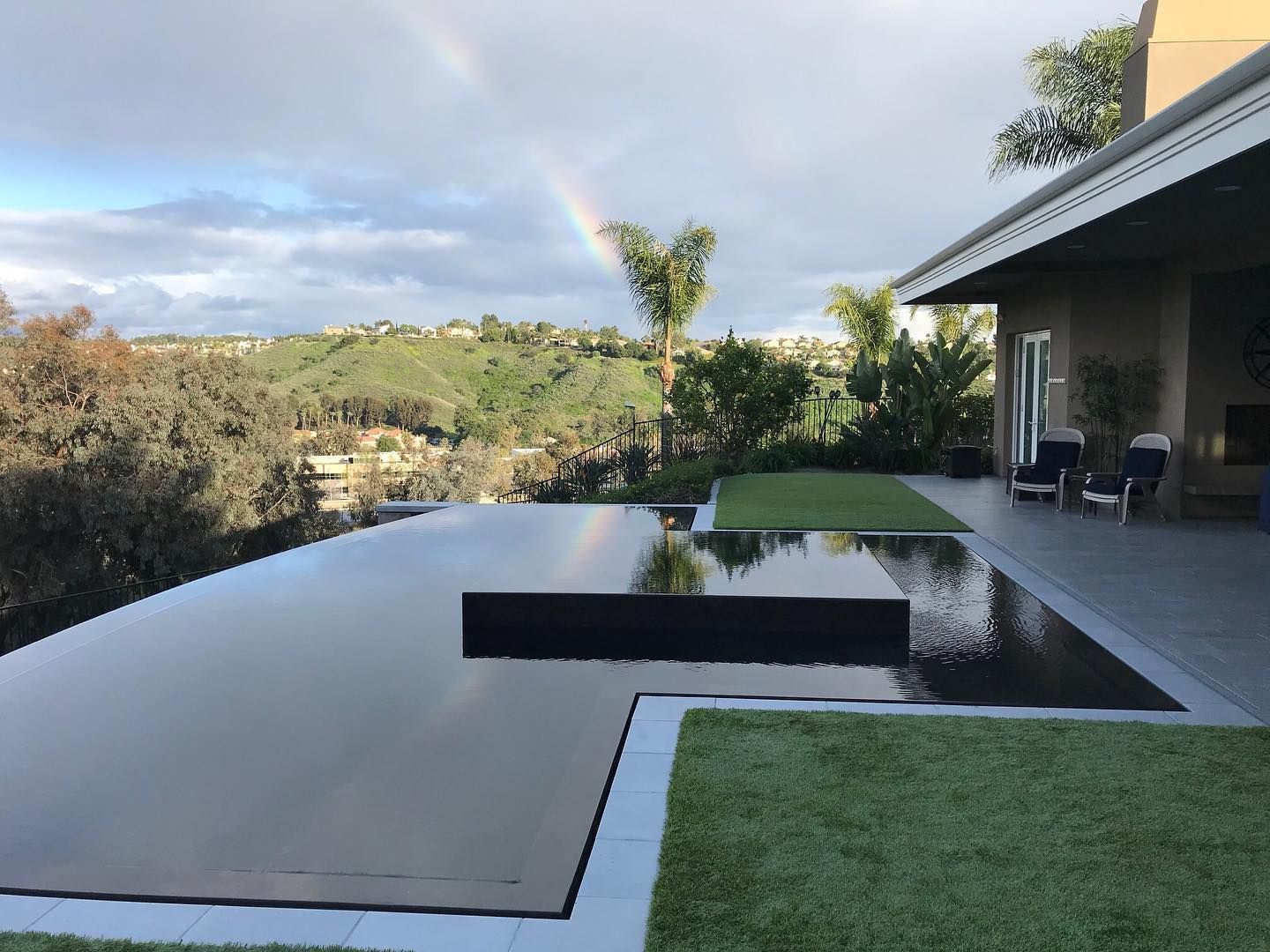 Infinity pool overlooking a valley with a rainbow. The pool reflects the cloudy sky. Adjacent is a lawn and a house.