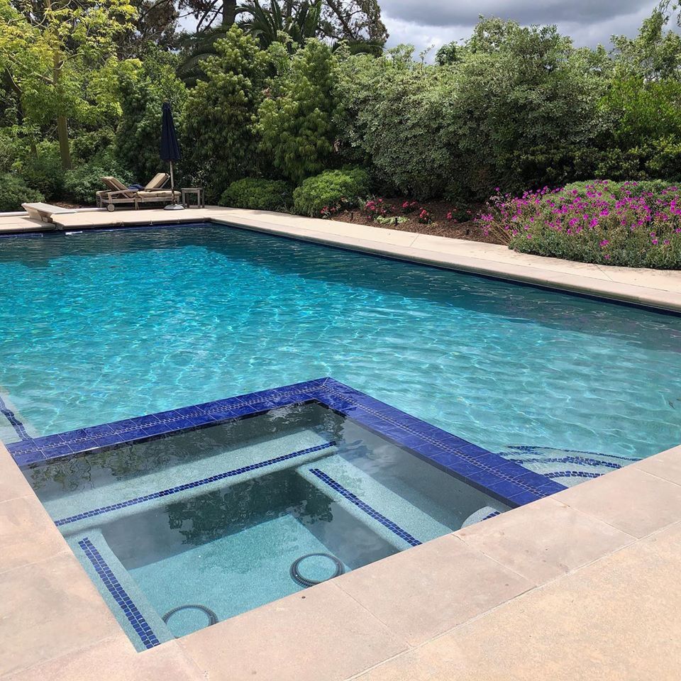 Swimming pool with attached spa, turquoise water, surrounded by tan stone and lush greenery.
