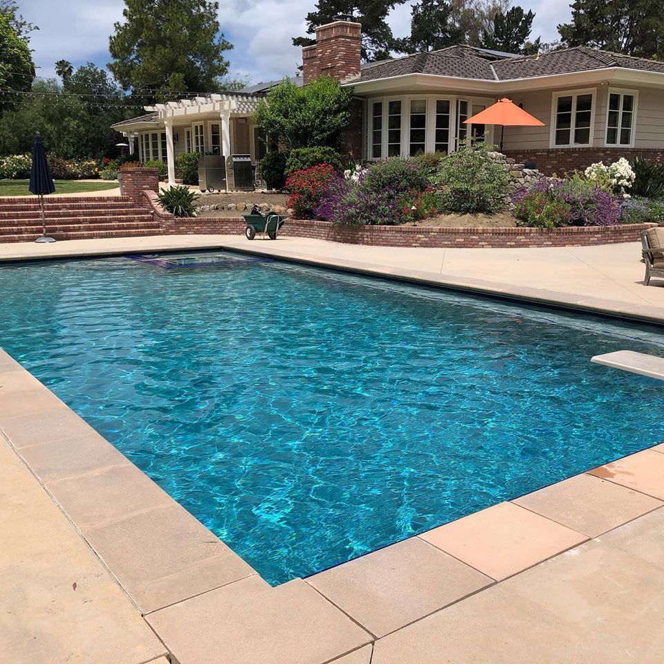 Swimming pool in front of a house. Turquoise water, beige stone surround. House has a brick chimney, and garden.