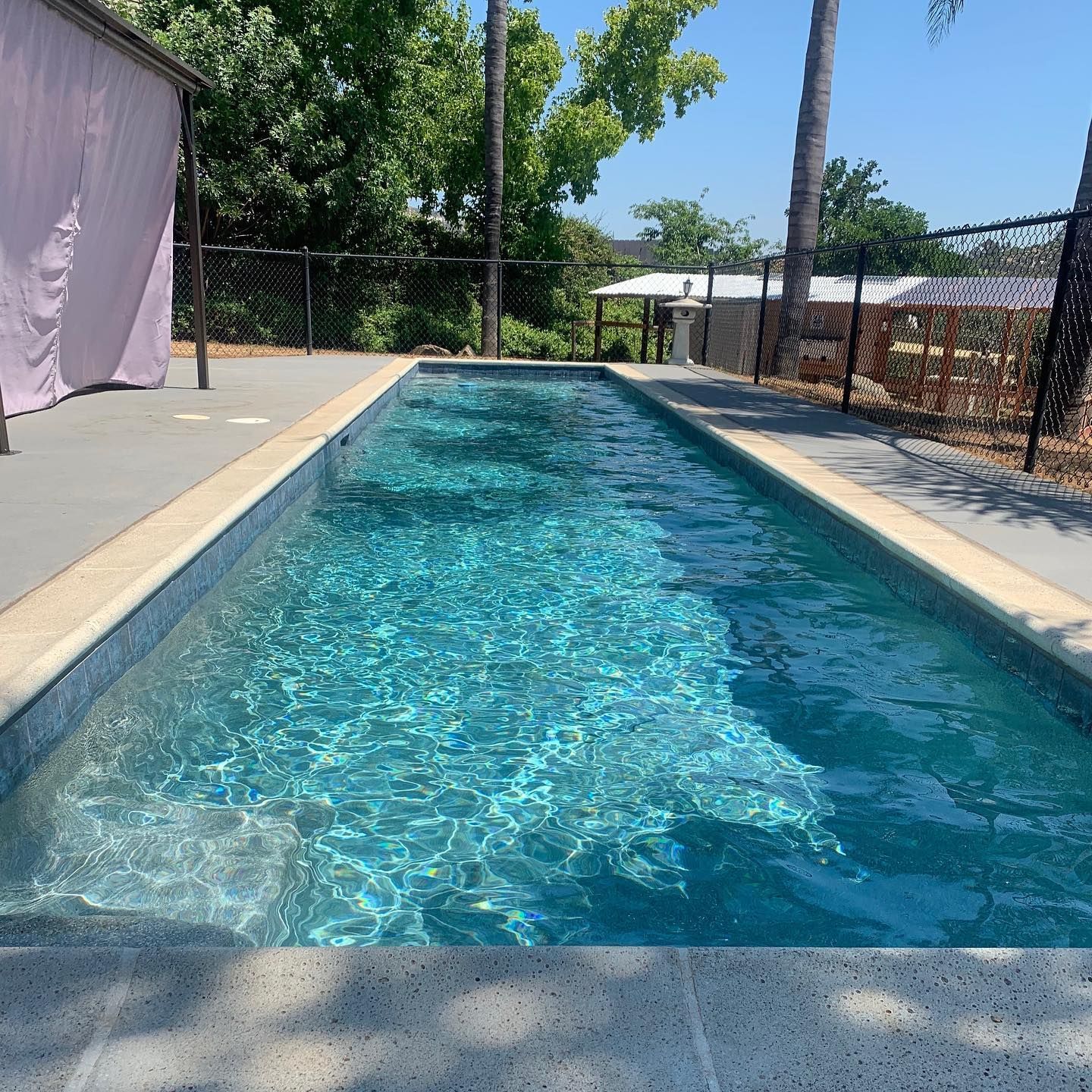 Rectangular outdoor pool with shimmering blue water, surrounded by concrete and a fence.