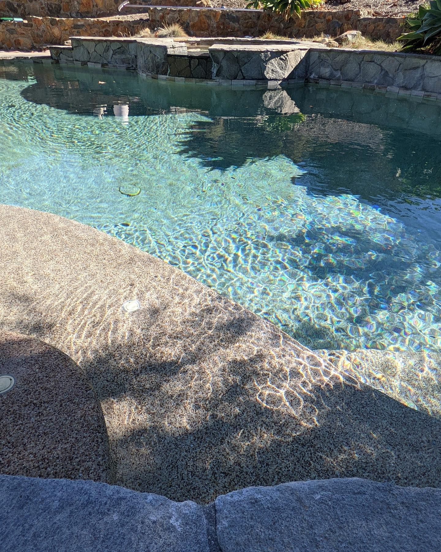 Clear, blue swimming pool with stone steps and tiered landscaping reflecting sunlight.
