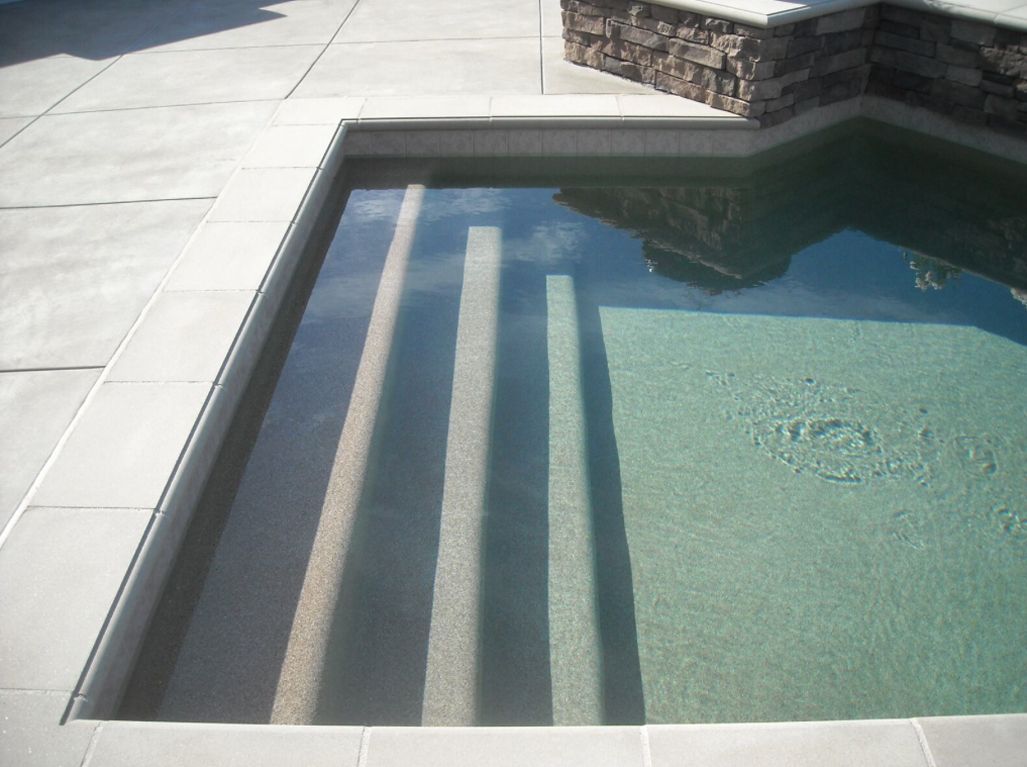 Swimming pool with steps leading into the water. Beige concrete surrounds the pool.