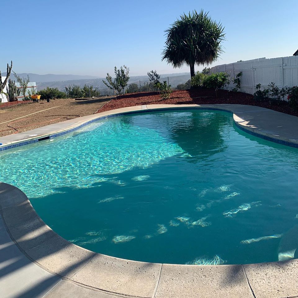 Pool with turquoise water on a sunny day, with a concrete border and a view of trees and mountains in the background.