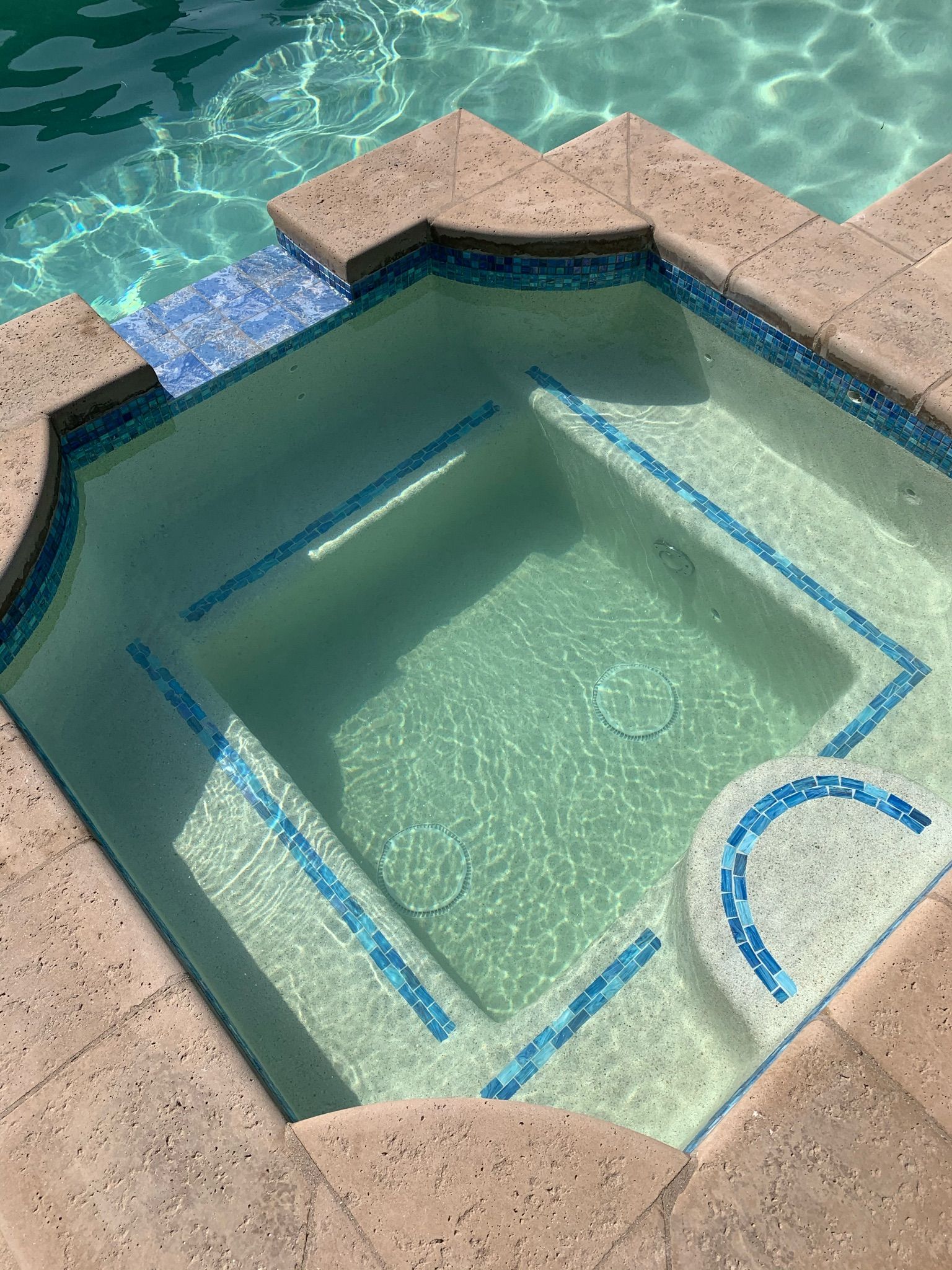 Small, square hot tub with blue tile trim and tan stone coping, filled with clear water.