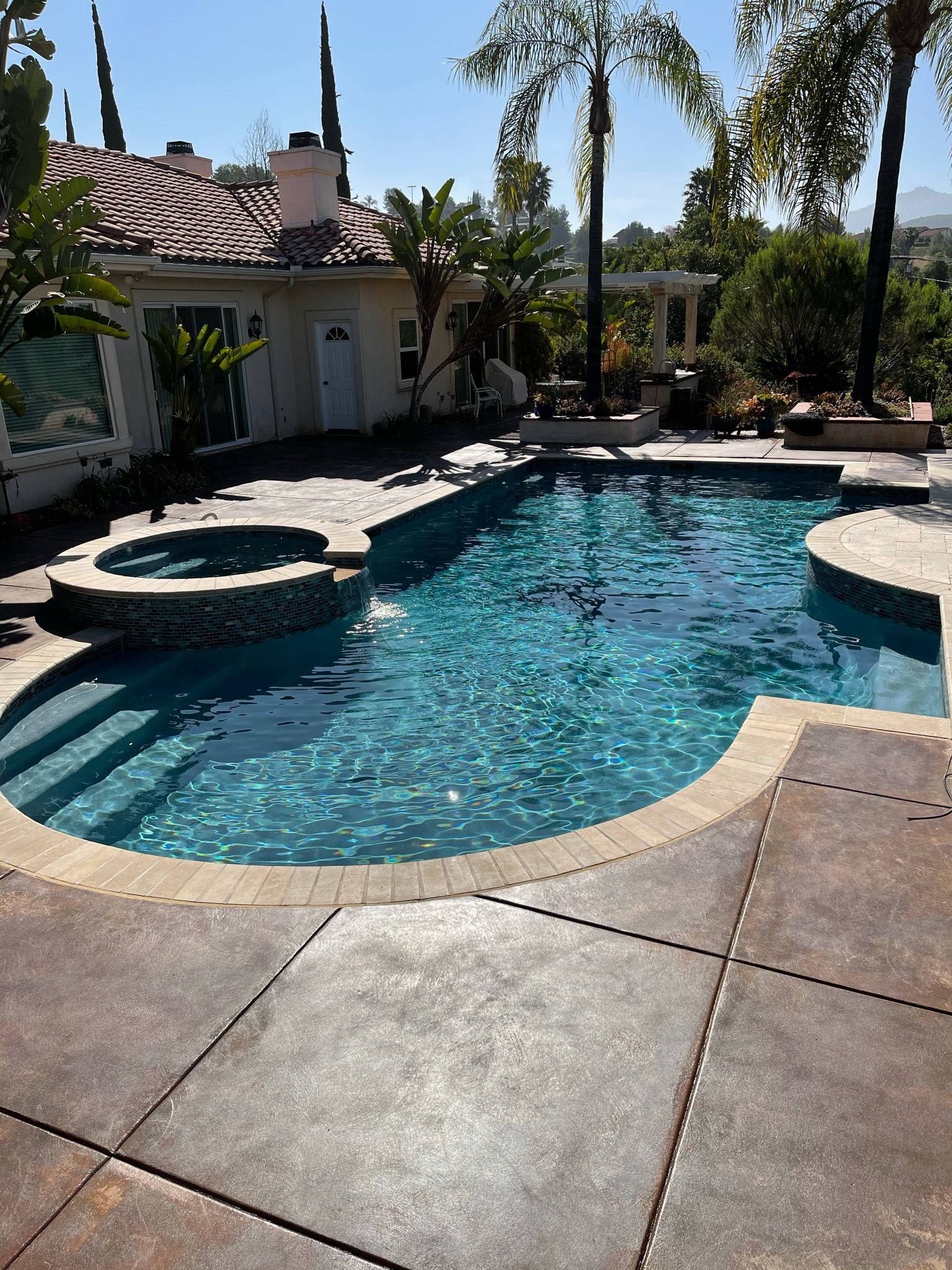 Pool with turquoise water, hot tub, and concrete patio in front of a house on a sunny day.