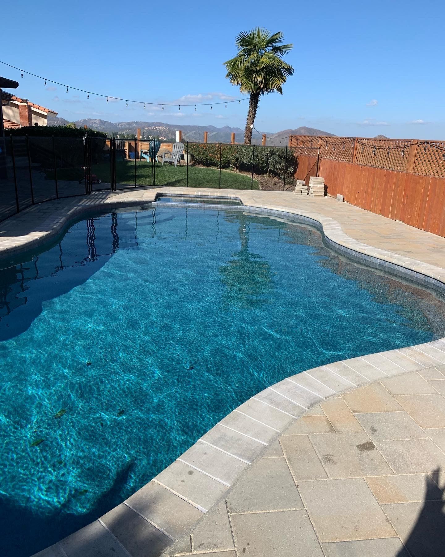 Pool with blue water and a stone patio, fence, tree, mountains, and string lights on a sunny day.