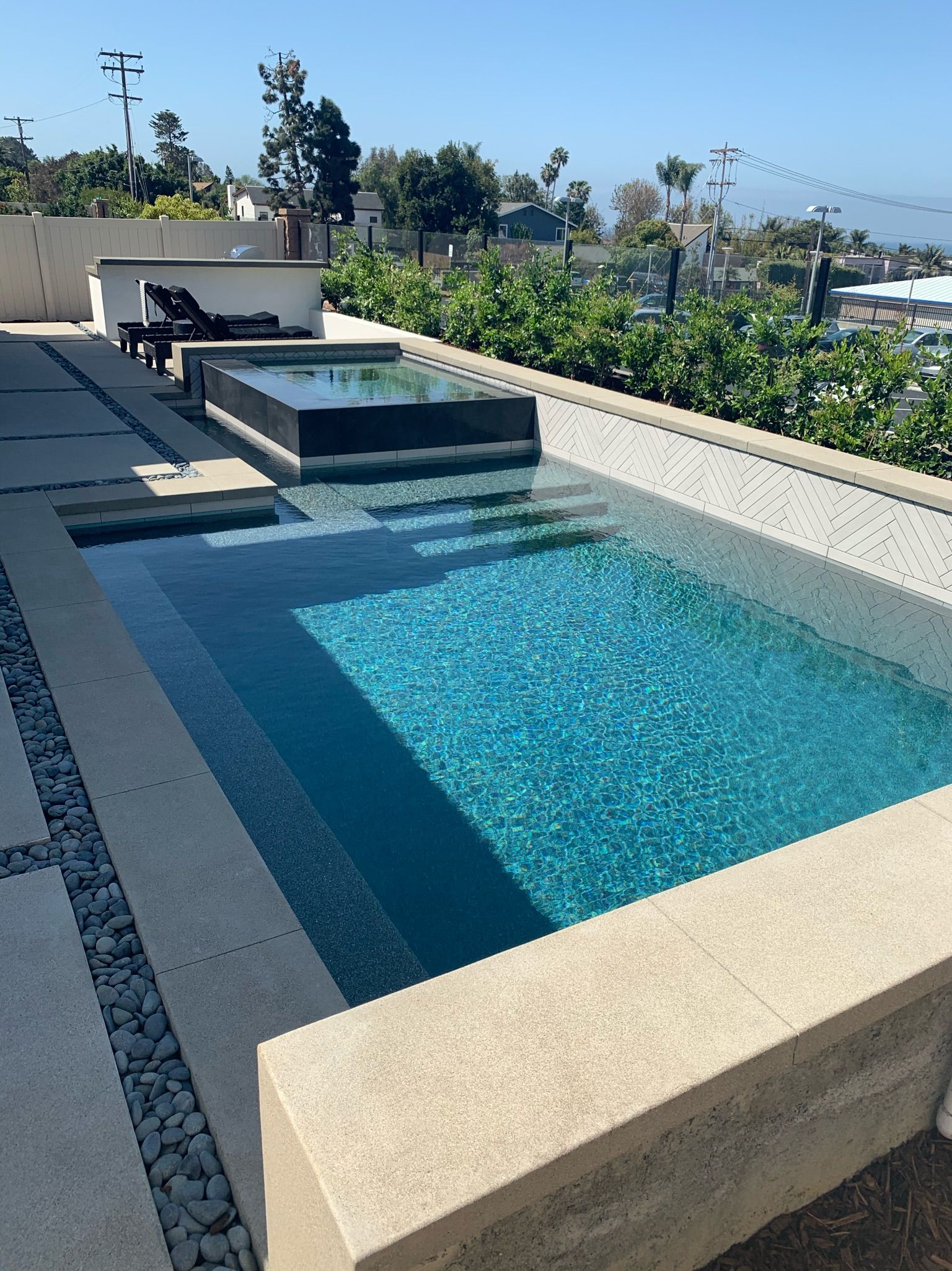 Rectangular swimming pool with a raised hot tub, stone patio, blue water, and greenery on a sunny day.