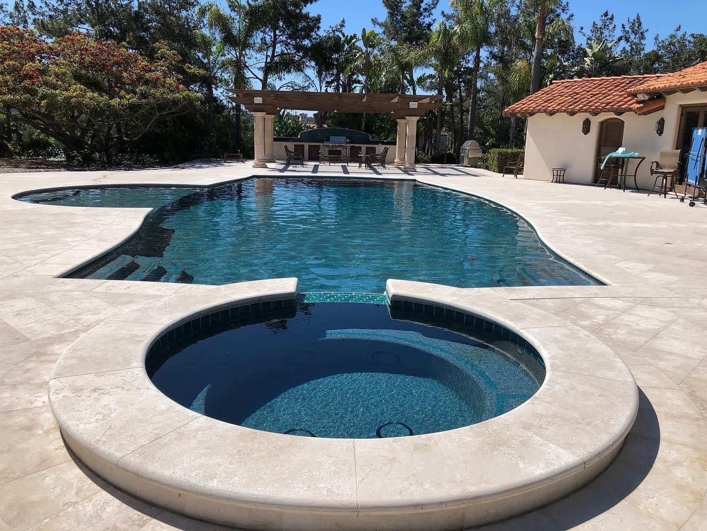 Swimming pool with attached jacuzzi, patio seating area, and a building under a blue sky.