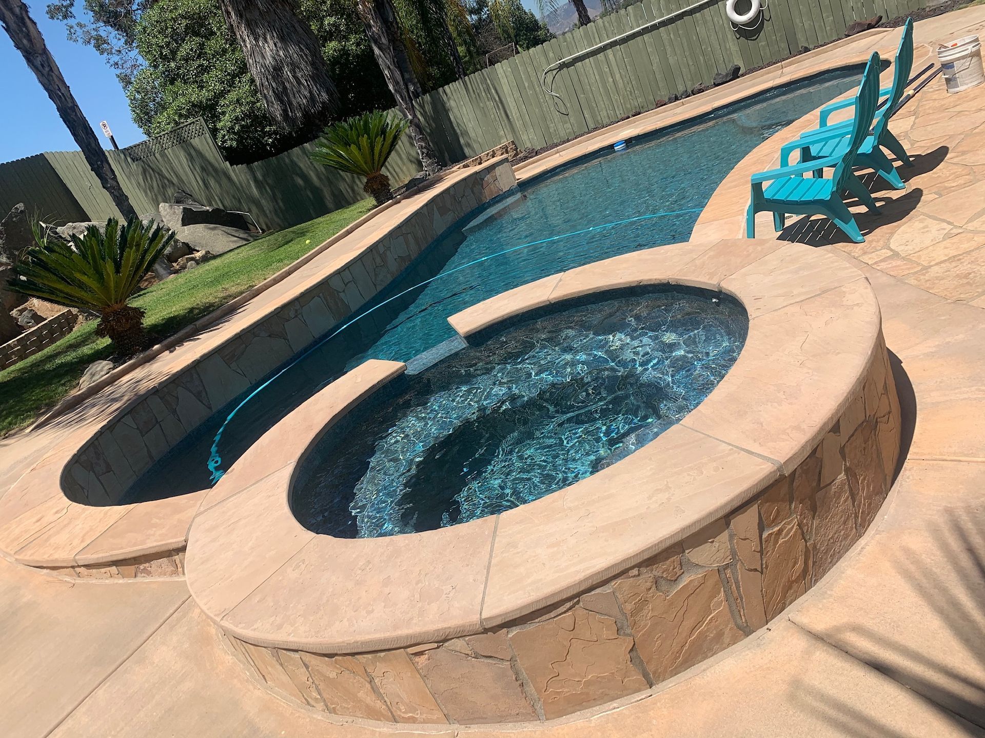 Pool and hot tub with turquoise chairs on a patio. Blue water reflects sunlight.