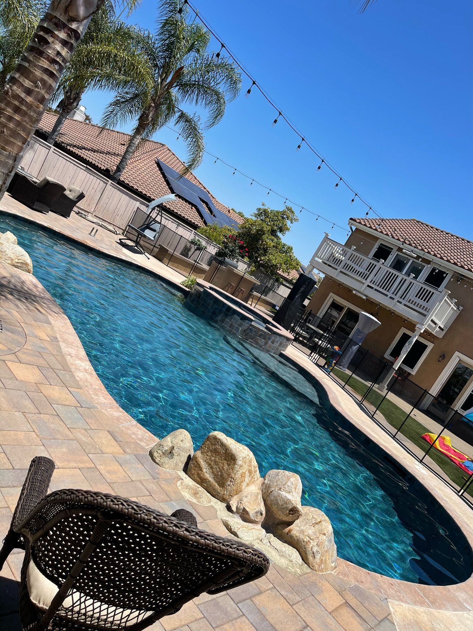 Poolside scene with a blue pool, a lounge chair, and a two-story house under a clear sky.