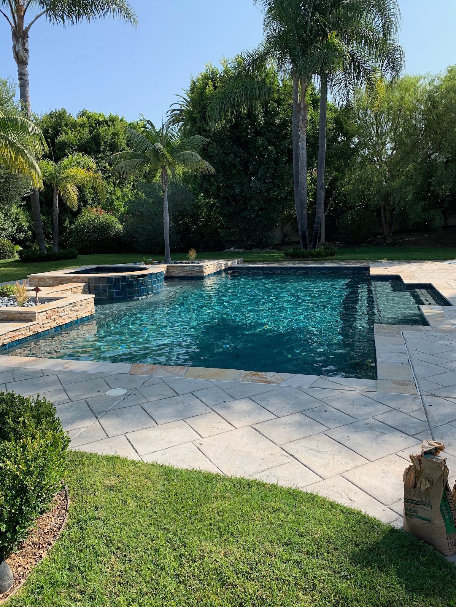 Pool with dark blue water surrounded by beige stone and green grass under a clear sky.