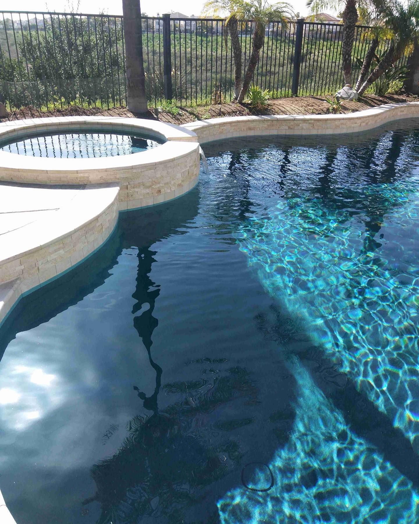 Pool with attached spa, blue water, stone coping, black fence in the background, sunny day.