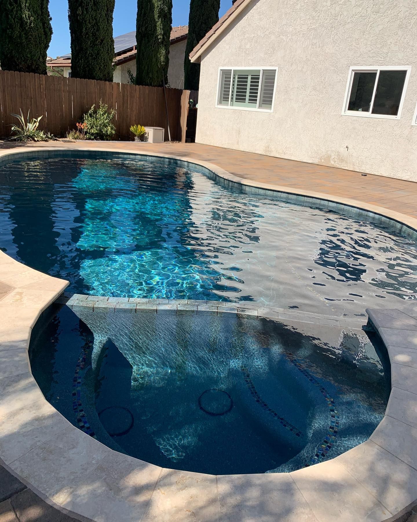 Pool with blue water and attached spa, surrounded by concrete and a house.