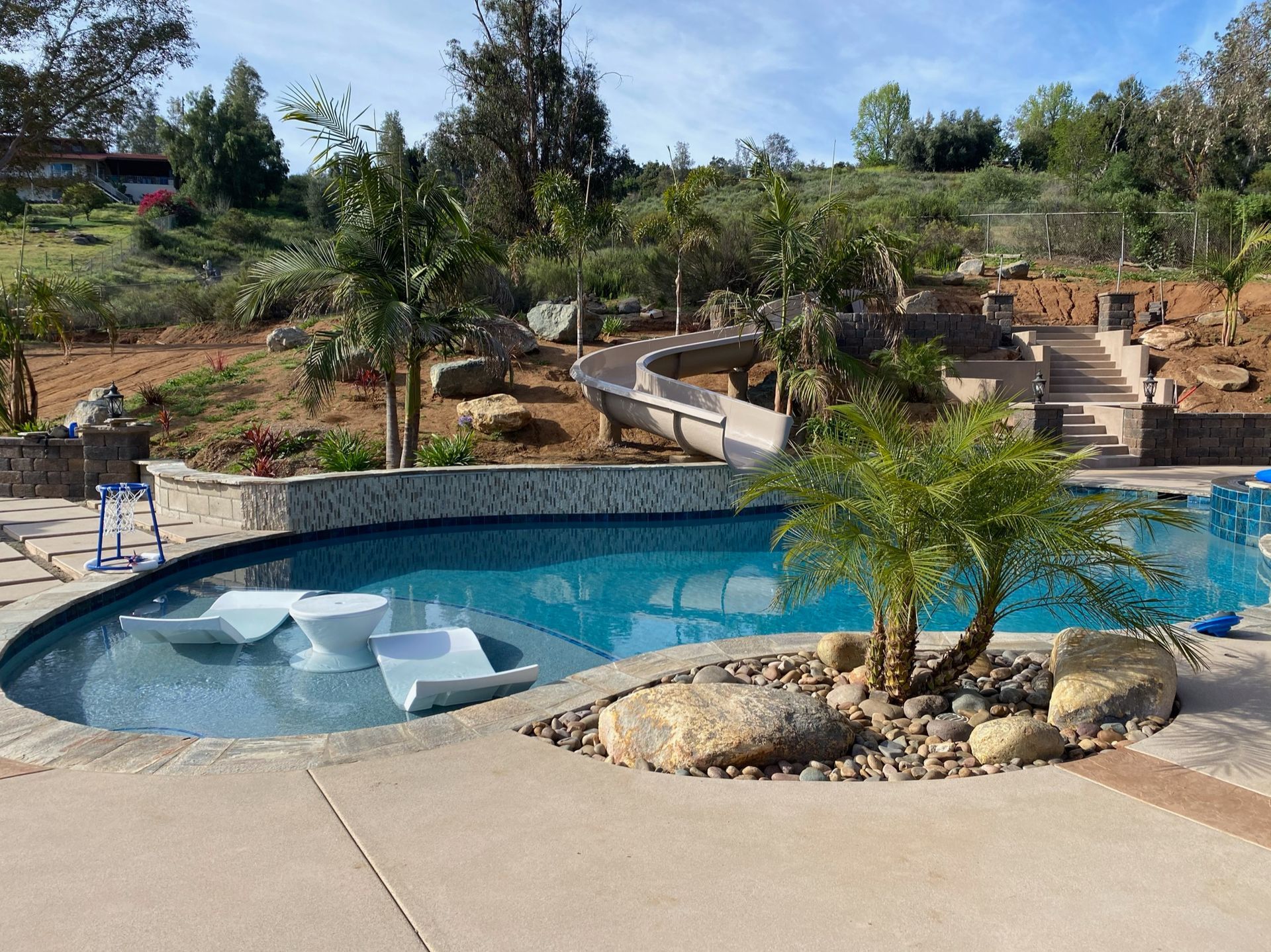 Swimming pool with built-in seating, palm trees, and a slide leading down from a hillside.