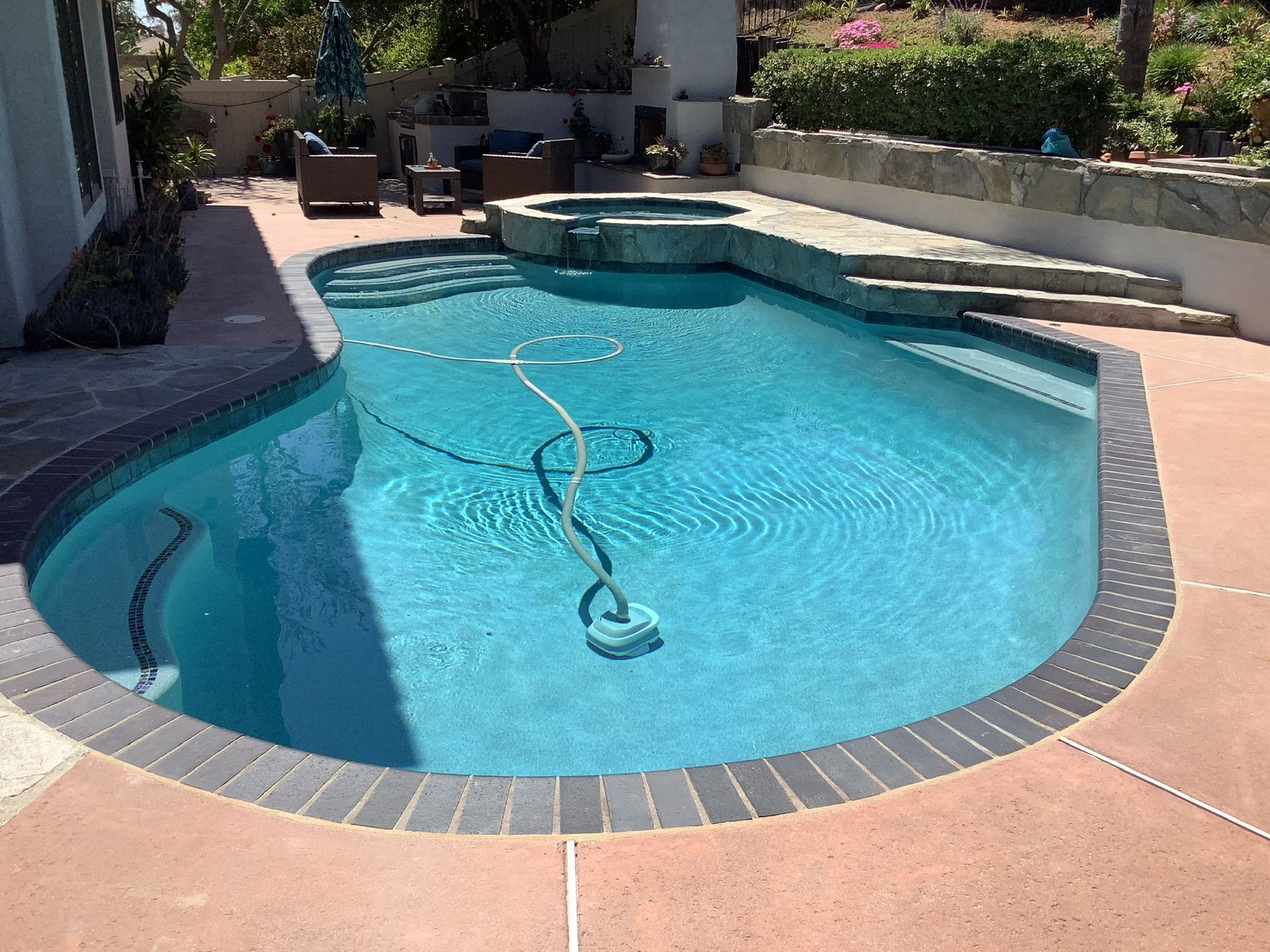 Swimming pool with blue water and pool cleaner, surrounded by a concrete deck and brick coping.