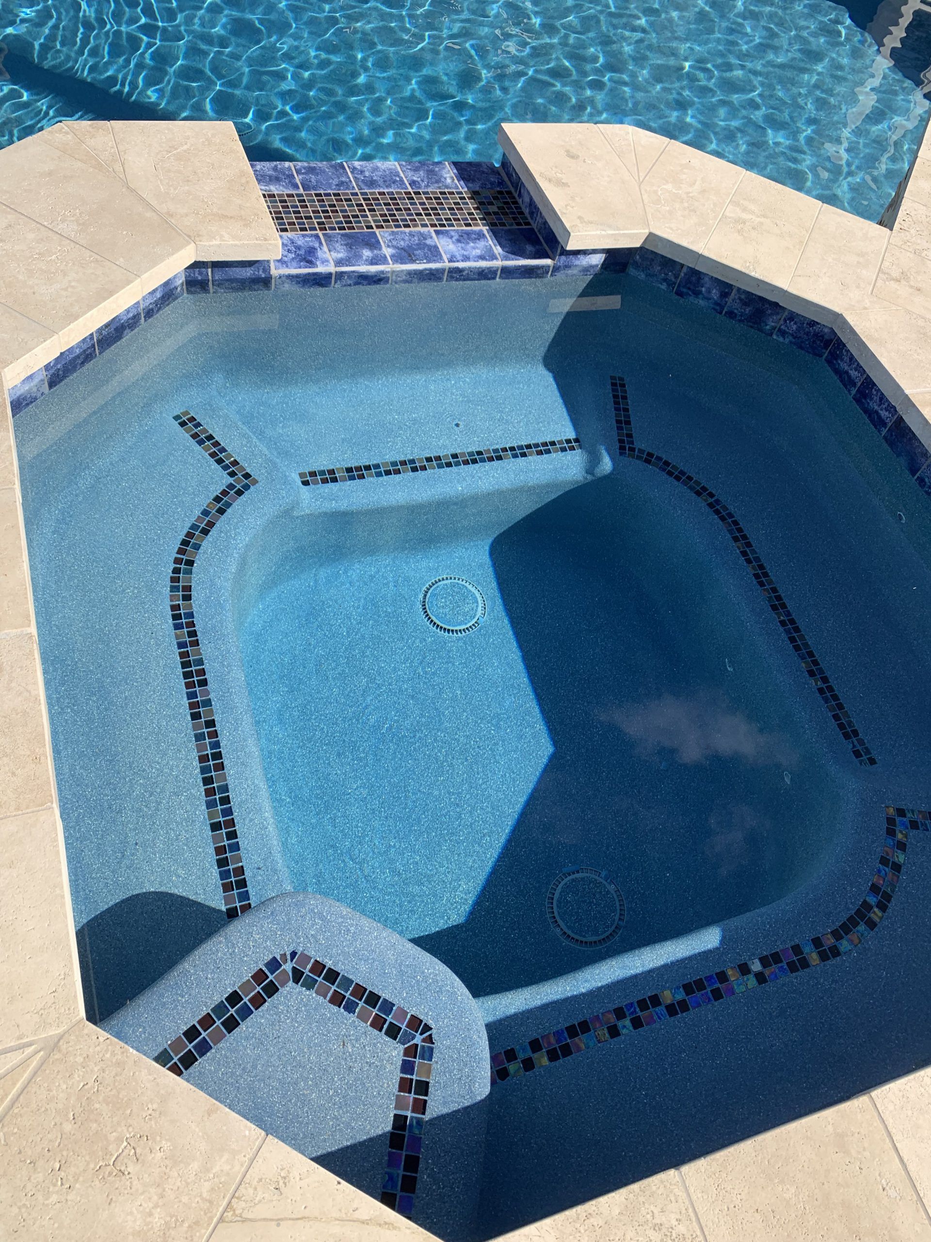 Blue-tiled jacuzzi with steps, connected to a larger pool. Beige stone border.