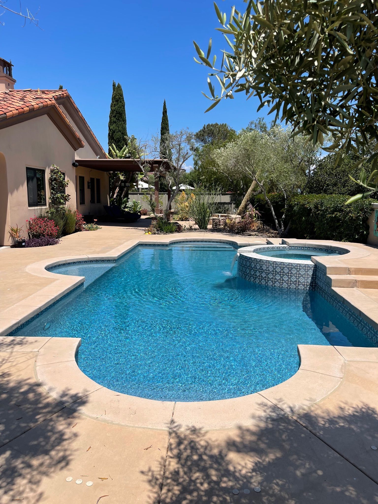 Swimming pool with blue water and adjacent spa, next to a house under a sunny sky.