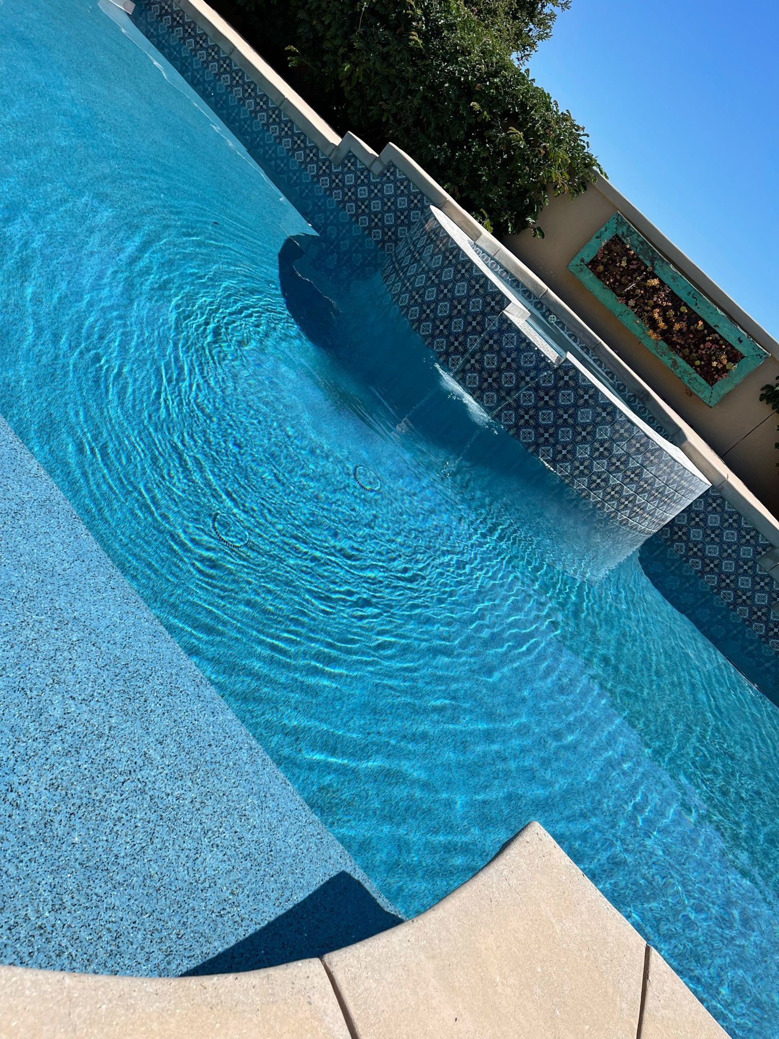 Blue tiled swimming pool with waterfall feature and steps, with a clear blue sky.