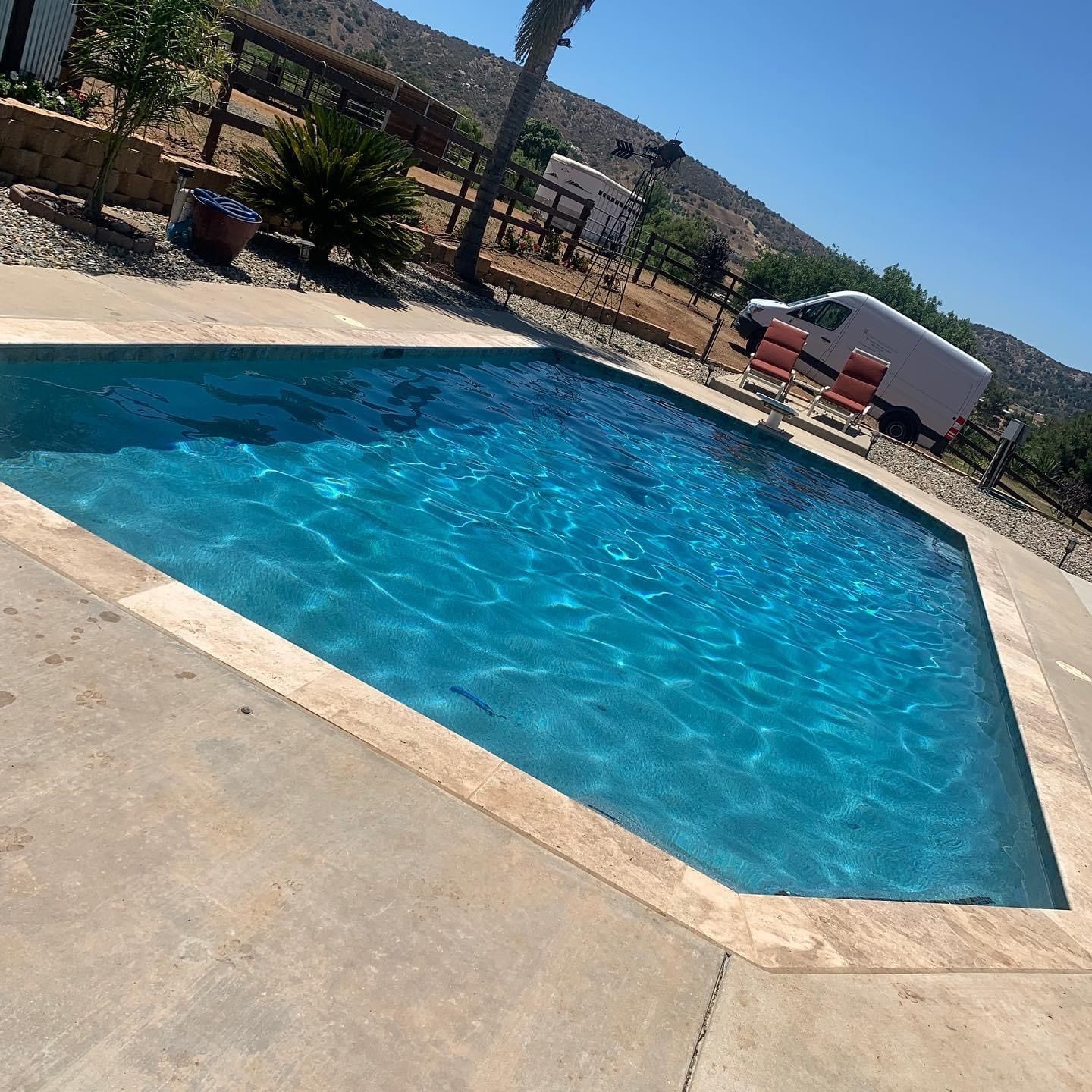 A blue pool with a stone surround, two red chairs, and a white van. Sunny outdoor setting.