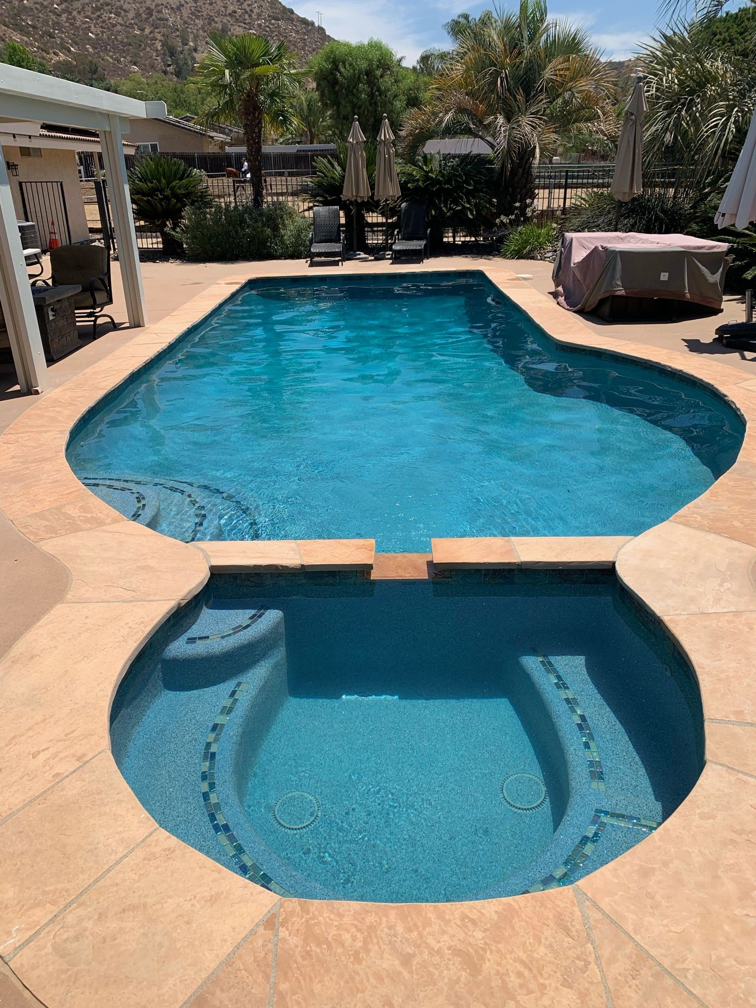 Pool with attached spa, surrounded by tan concrete. Blue water, sunny day, residential setting.