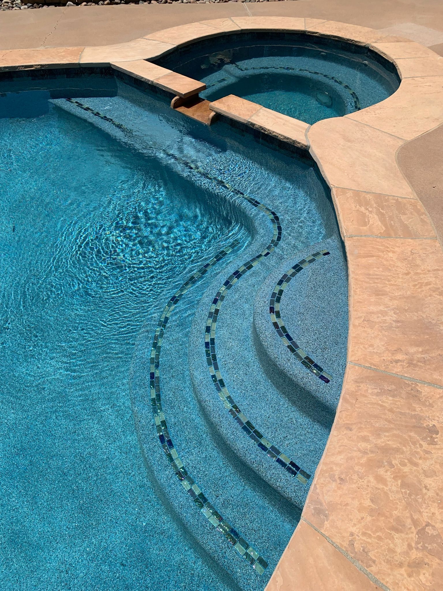 Swimming pool with curved steps and adjacent jacuzzi, blue water, and tan stone edging.