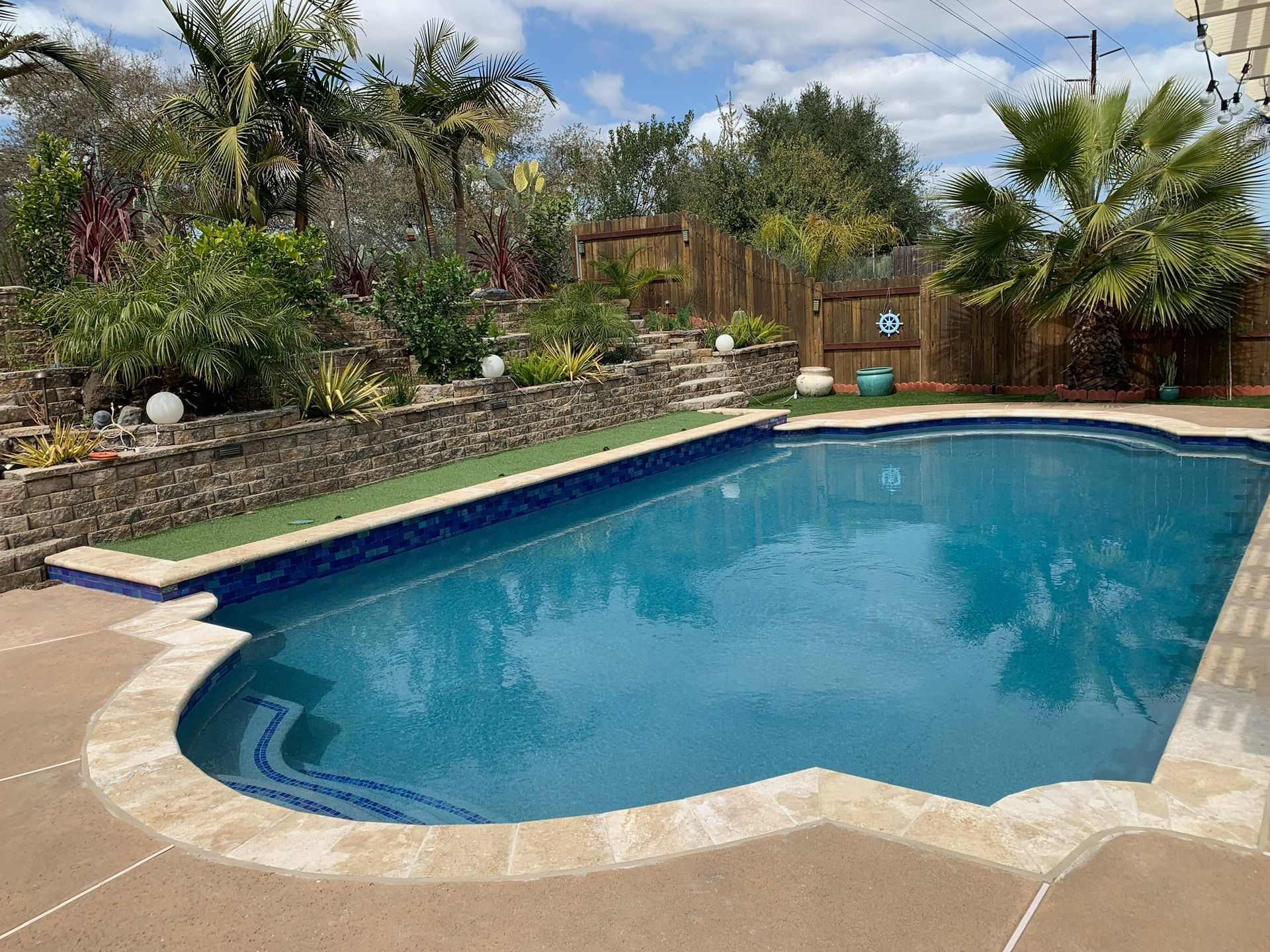 Swimming pool surrounded by landscaping with palm trees and a wooden fence, under a partly cloudy sky.