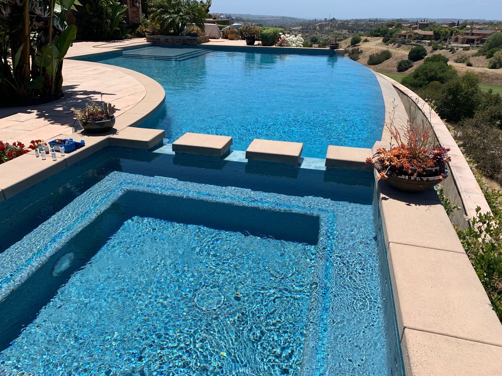 Pool with infinity edge overlooking a landscape, steps, and spa. Blue water, sunny day.