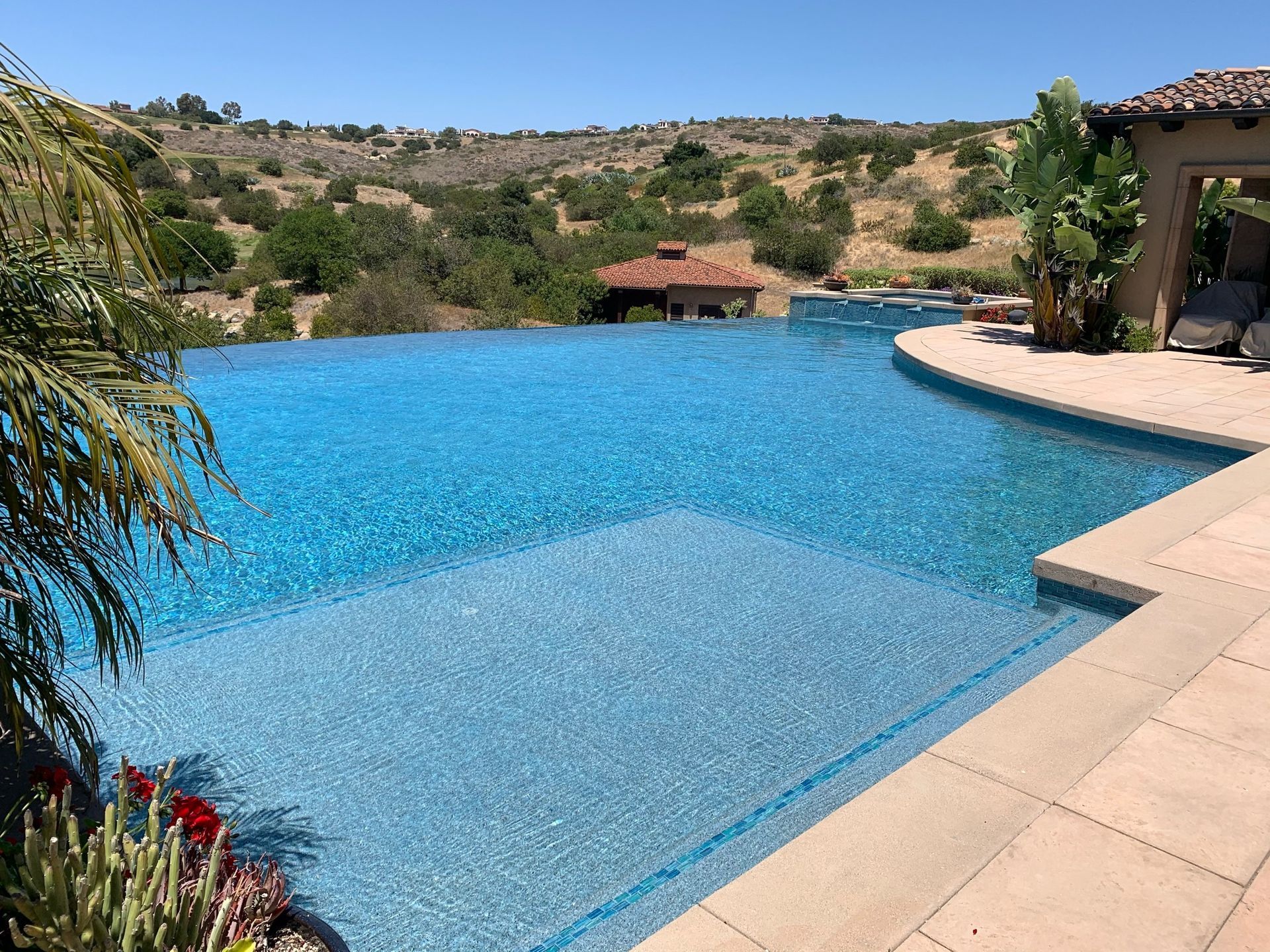 Infinity pool overlooking a landscape; blue water, sunny day, beige tile, lush greenery, and rolling hills in the background.