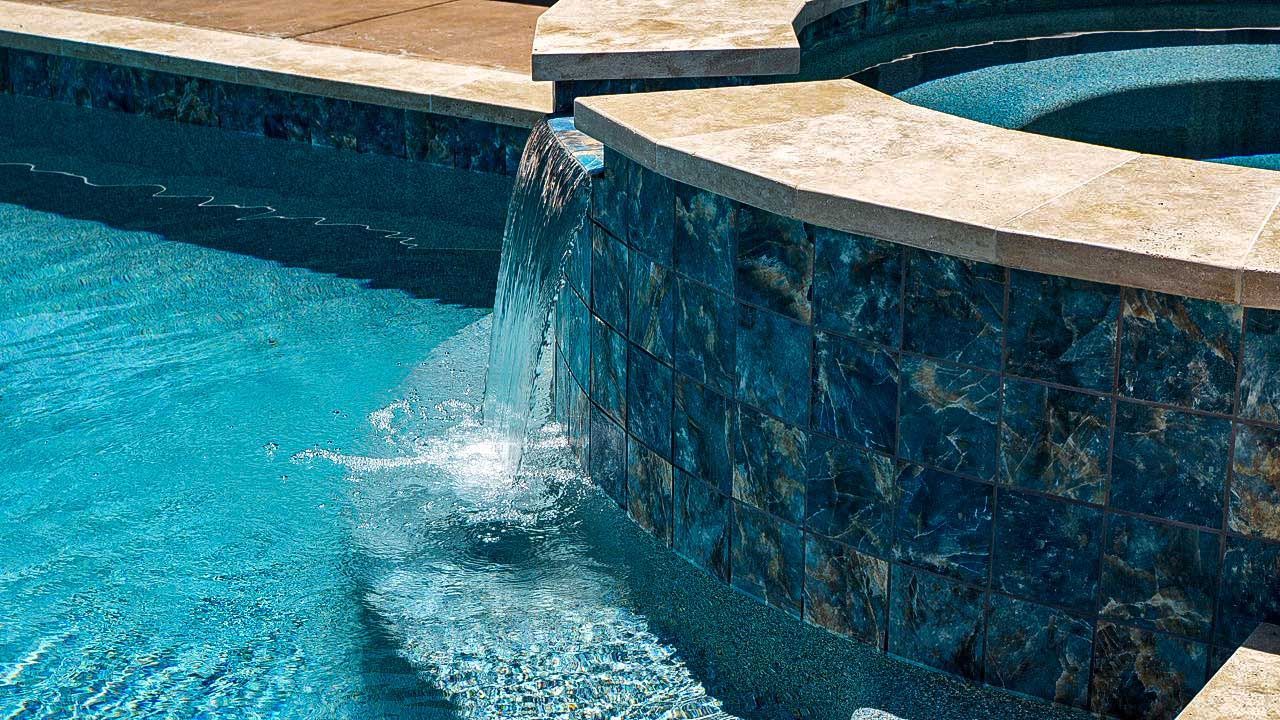 Pool with waterfall cascading over a dark blue tiled wall, flowing into the clear blue water.