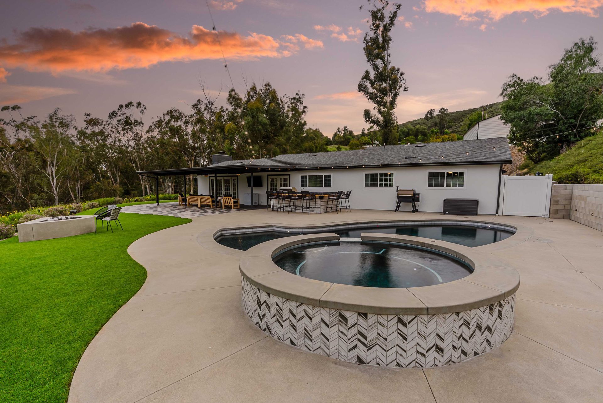 Backyard with pool and spa, white house, and green lawn at sunset.