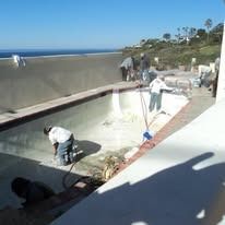Workers cleaning a large, empty swimming pool near the ocean.