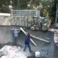 Workers constructing a pool with stone features, using wood beams, concrete, and tools.