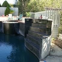Pool with dark tile, steps, and surrounding deck. Trees and greenery in background.